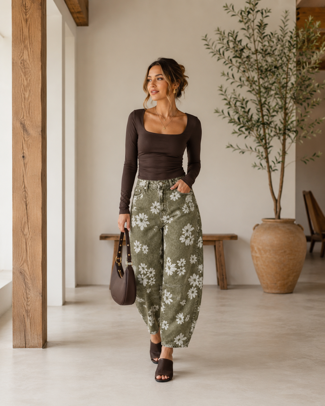 Woman wearing floral olive barrel jeans with a fitted chocolate brown square neck top, brown heeled sandals, layered gold jewelry, and a matching brown shoulder bag in a bright neutral luxury home with warm natural lighting.