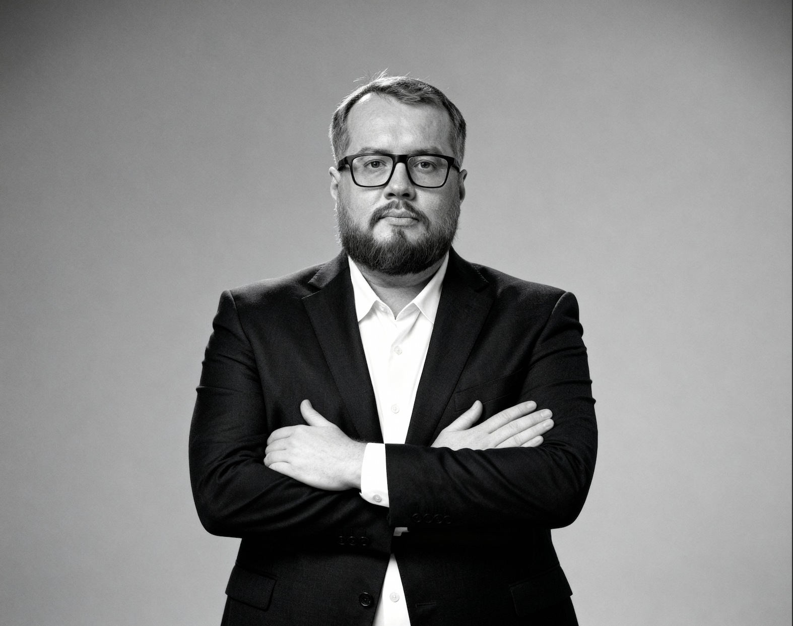 Black and white portrait of a man with glasses and a beard, wearing a suit and white shirt, crossing his arms.