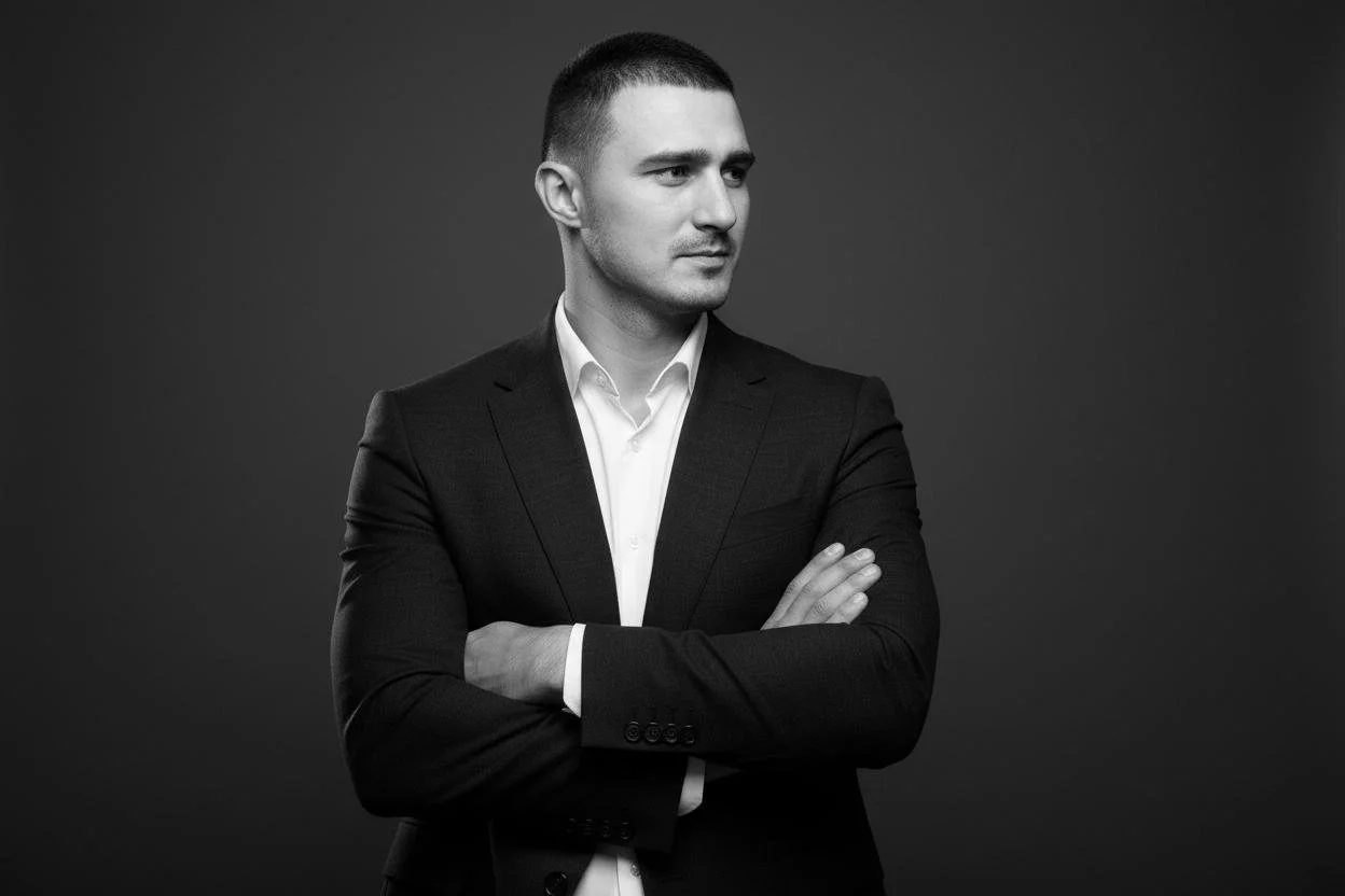 Black and white portrait of a man with short hair, wearing a dark suit and white shirt, standing with arms crossed against a plain background.