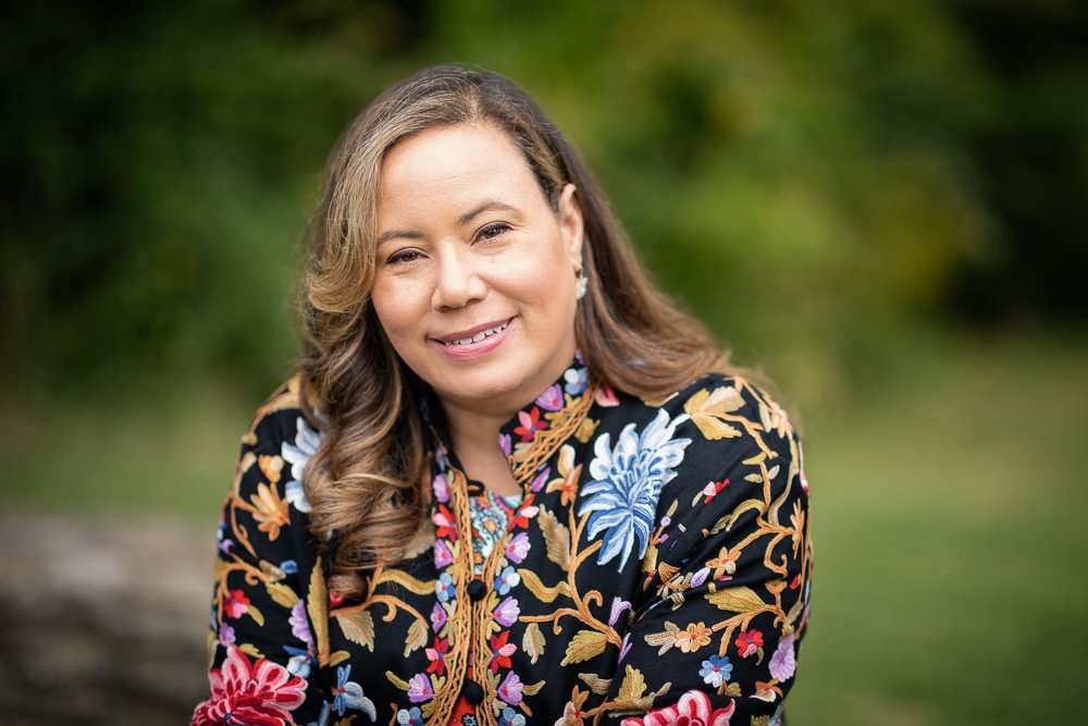 A woman with light skin, brown hair styled in loose curls, and wearing earrings, smiling outdoors in front of greenery, dressed in a black floral embroidered jacket.