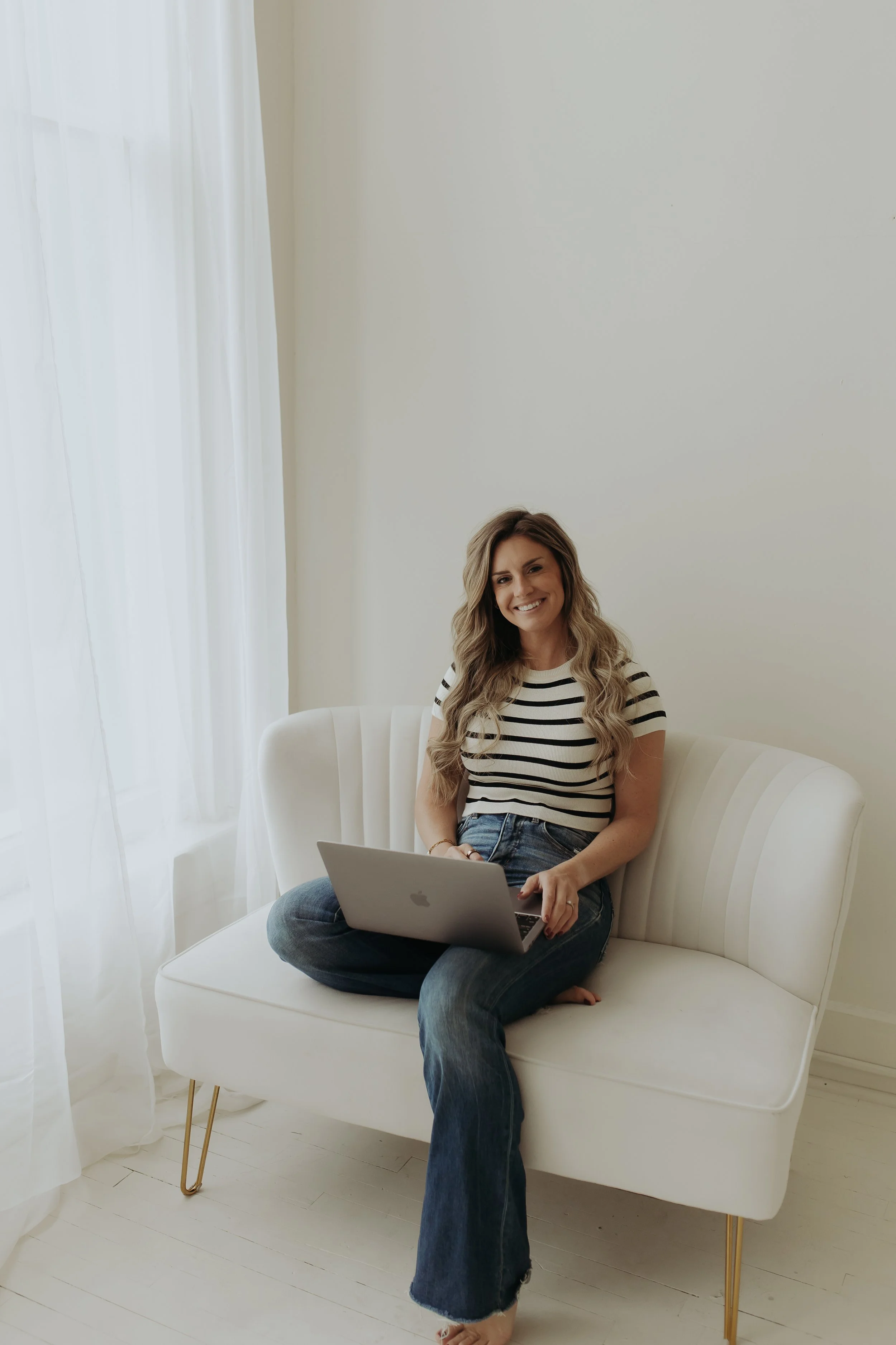 A woman sitting cross-legged on a white couch with a laptop on her lap, smiling, in a bright room with white walls and curtains.