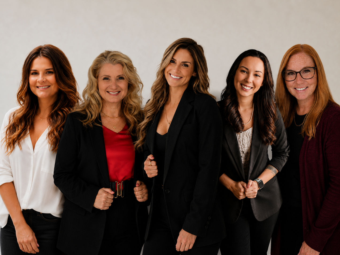 Group of five diverse women standing together, smiling, dressed in business casual attire against a plain background.