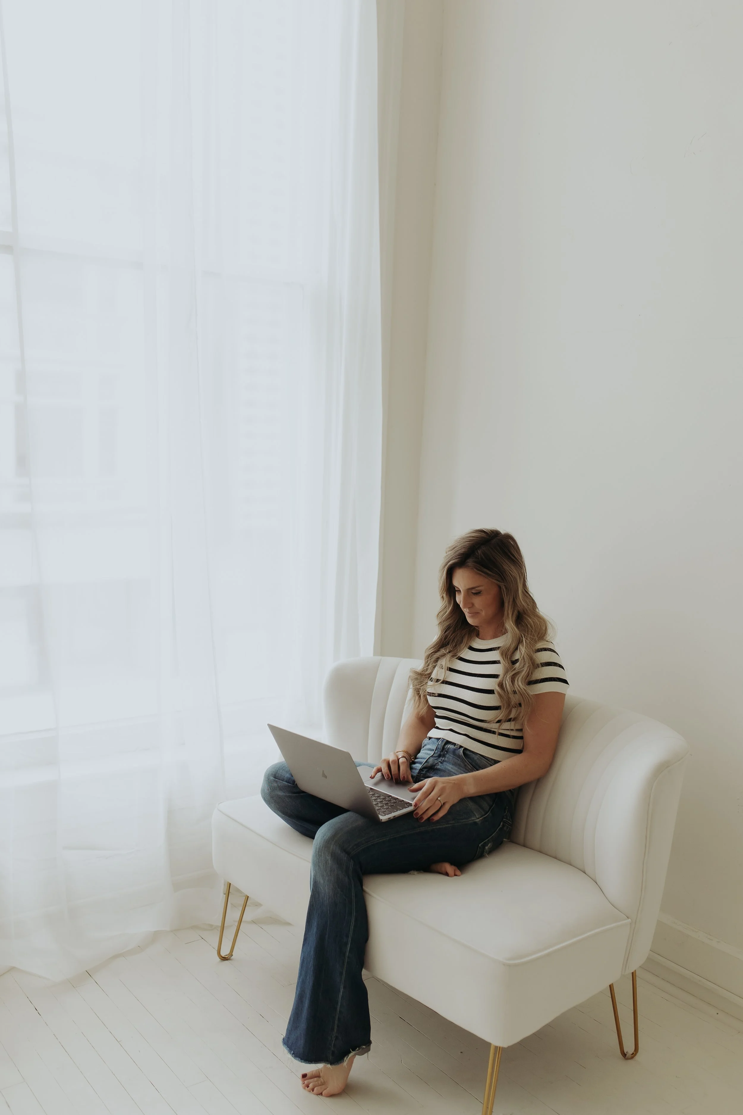 A woman sitting on a white sofa using a laptop, with a bright window and sheer curtains in the background.