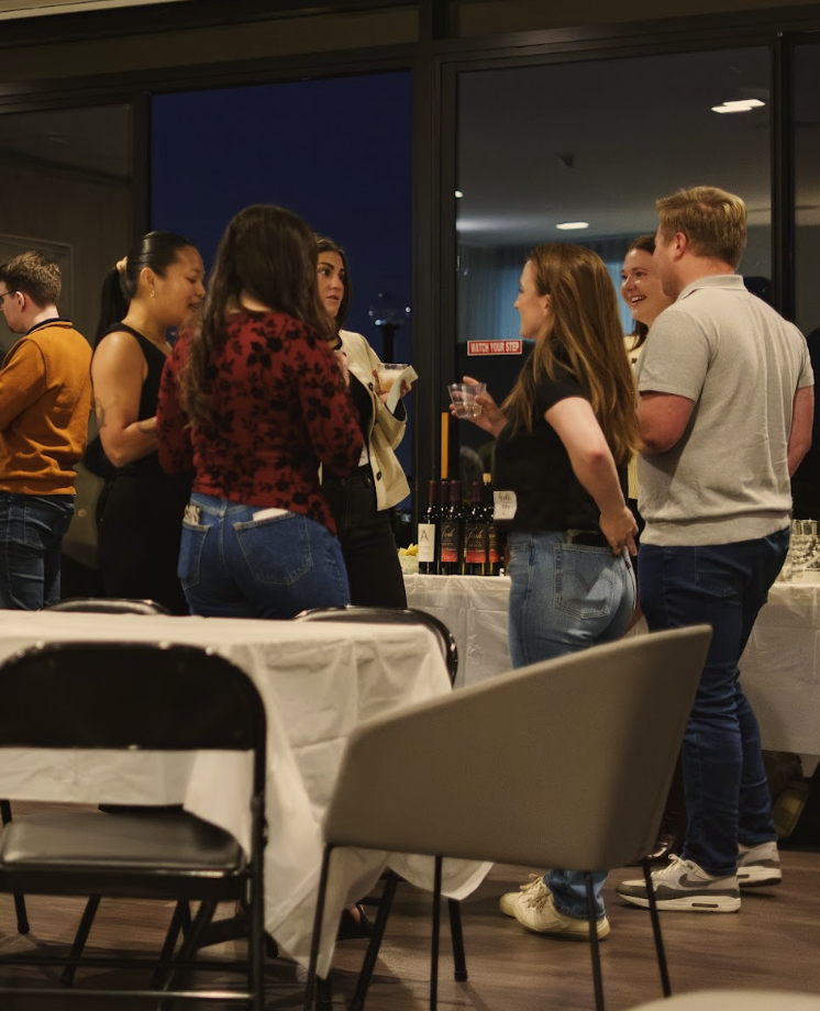 Group of people socializing at a table with wine bottles at an indoor event during the evening.