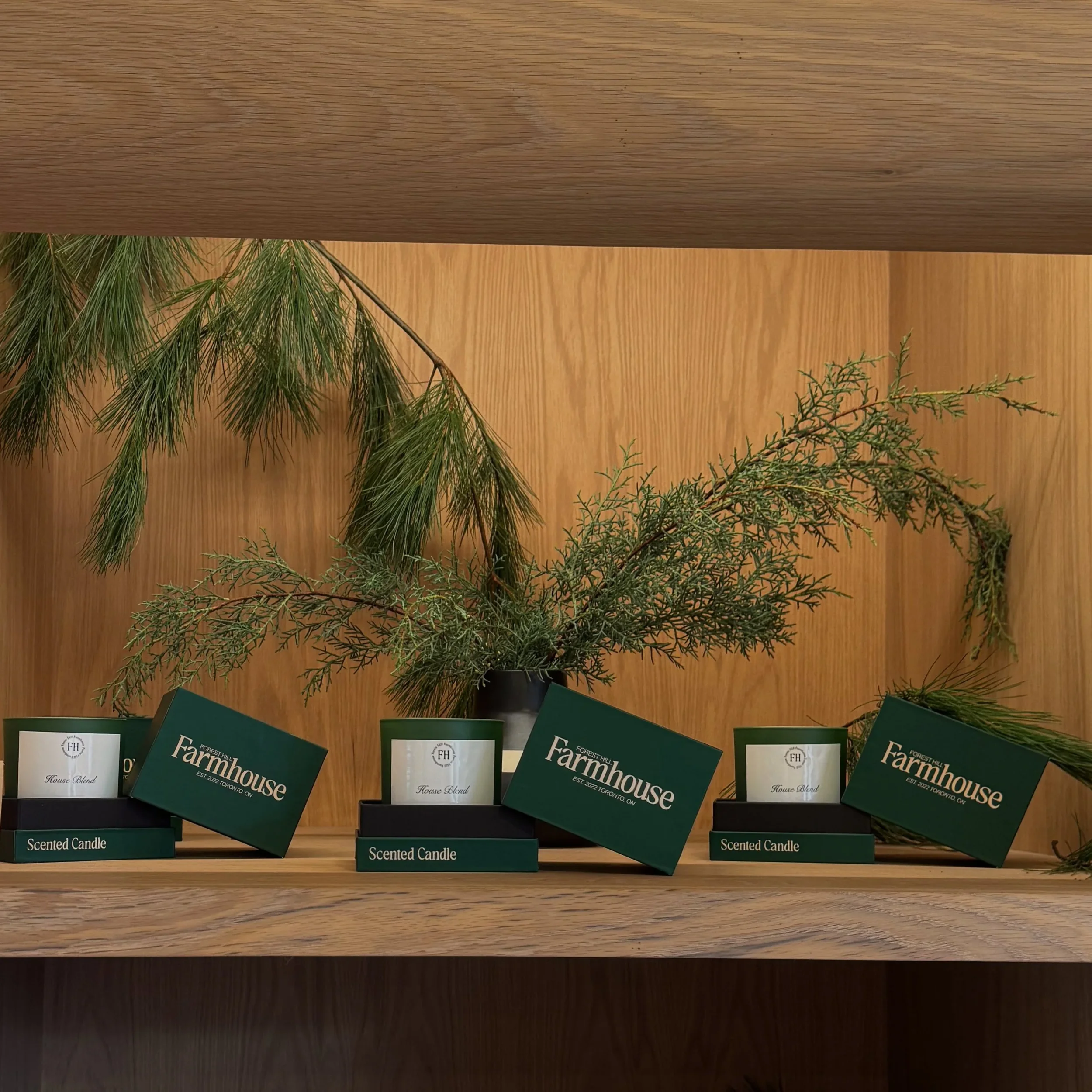 Display of scented candles with green farmhouse labels, arranged with pine branches on a wooden shelf.