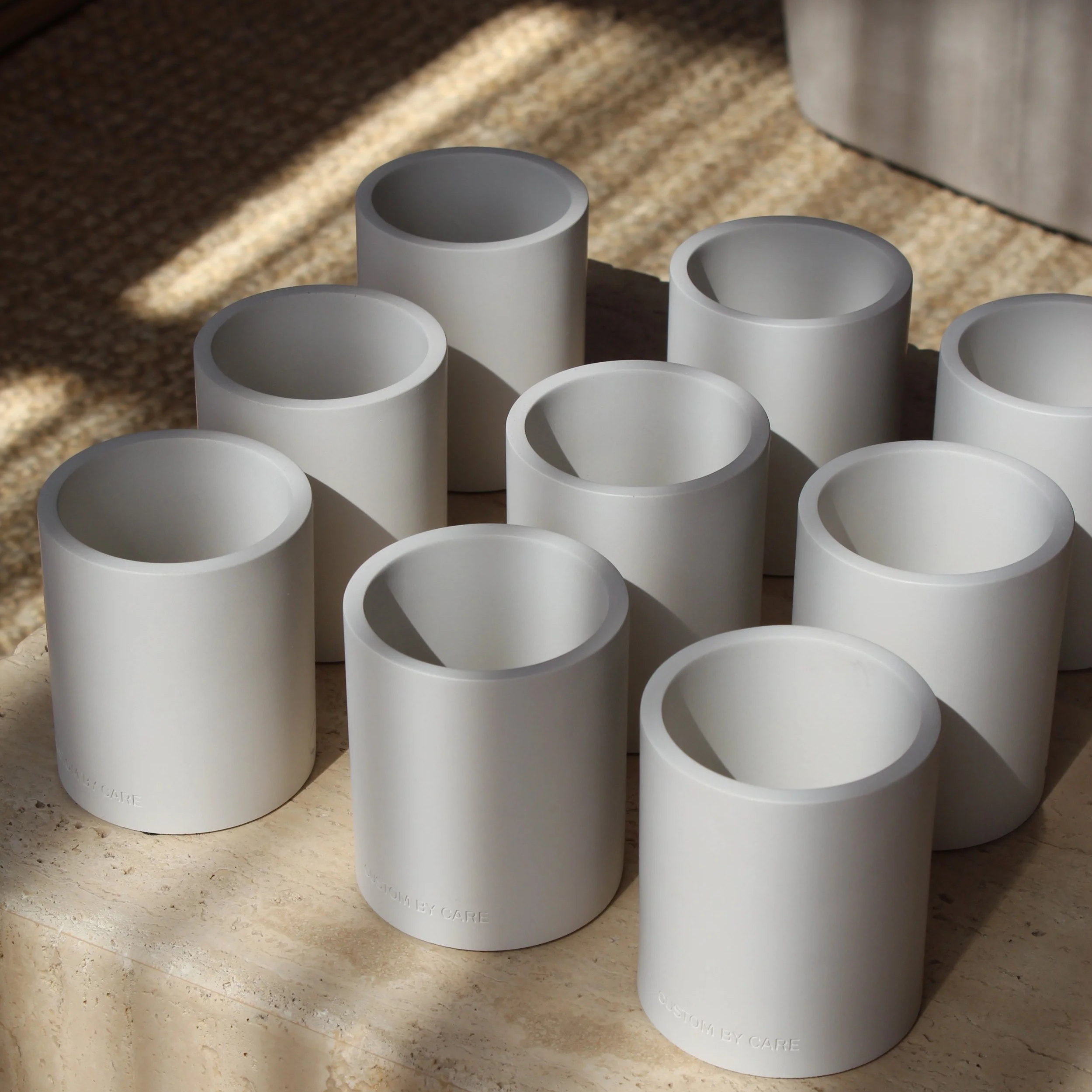Nine white cylindrical ceramic or porcelain cups arranged on a beige surface, with shadows cast by sunlight.
