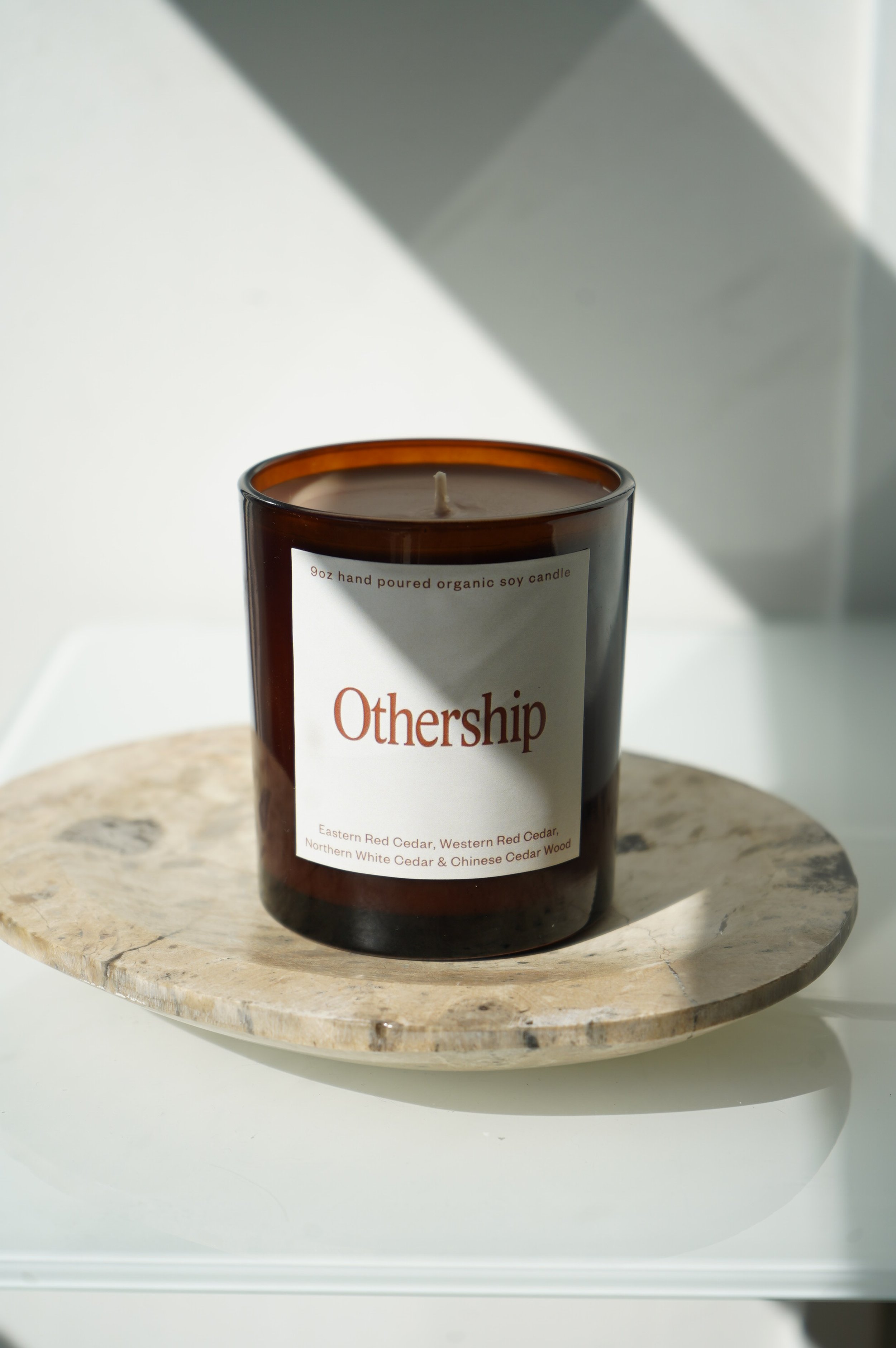 A brown candle jar labeled "Othership" placed on a round stone surface with sunlight casting shadows.