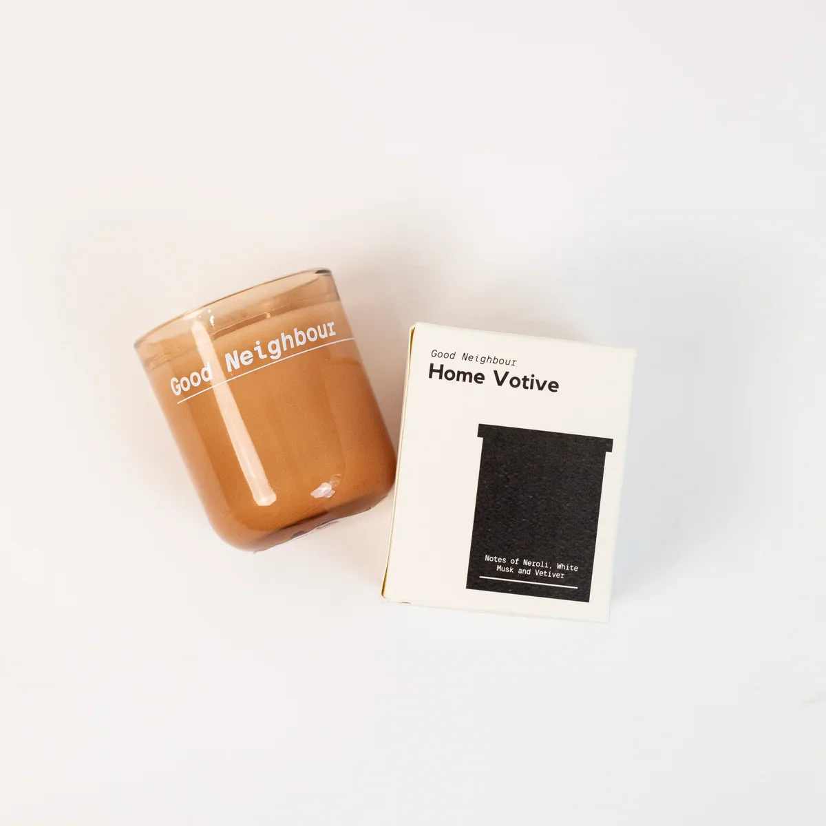 A brown candle with white text that says "Good Neighbour" next to a white box labeled "Good Neighbour Home Votive," featuring a black silhouette of a candle and notes about Neroli, White Musk, and Vetiver scents.