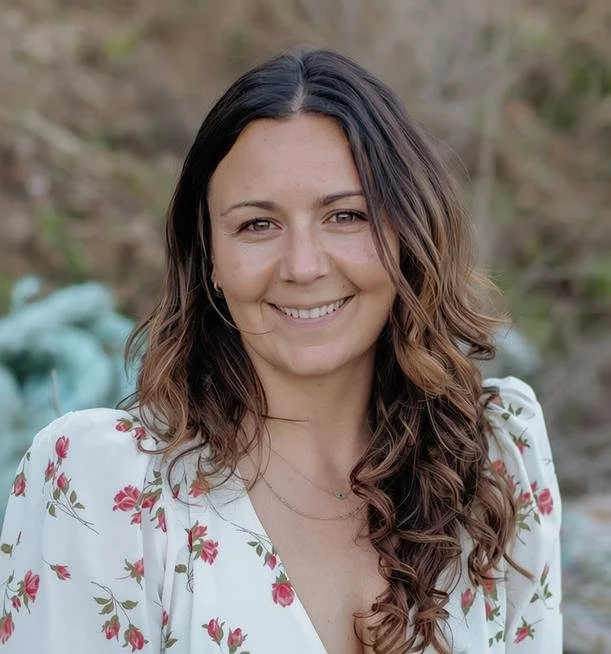 A woman with long, wavy brown hair wearing a white floral blouse outdoors with a blurred natural background.