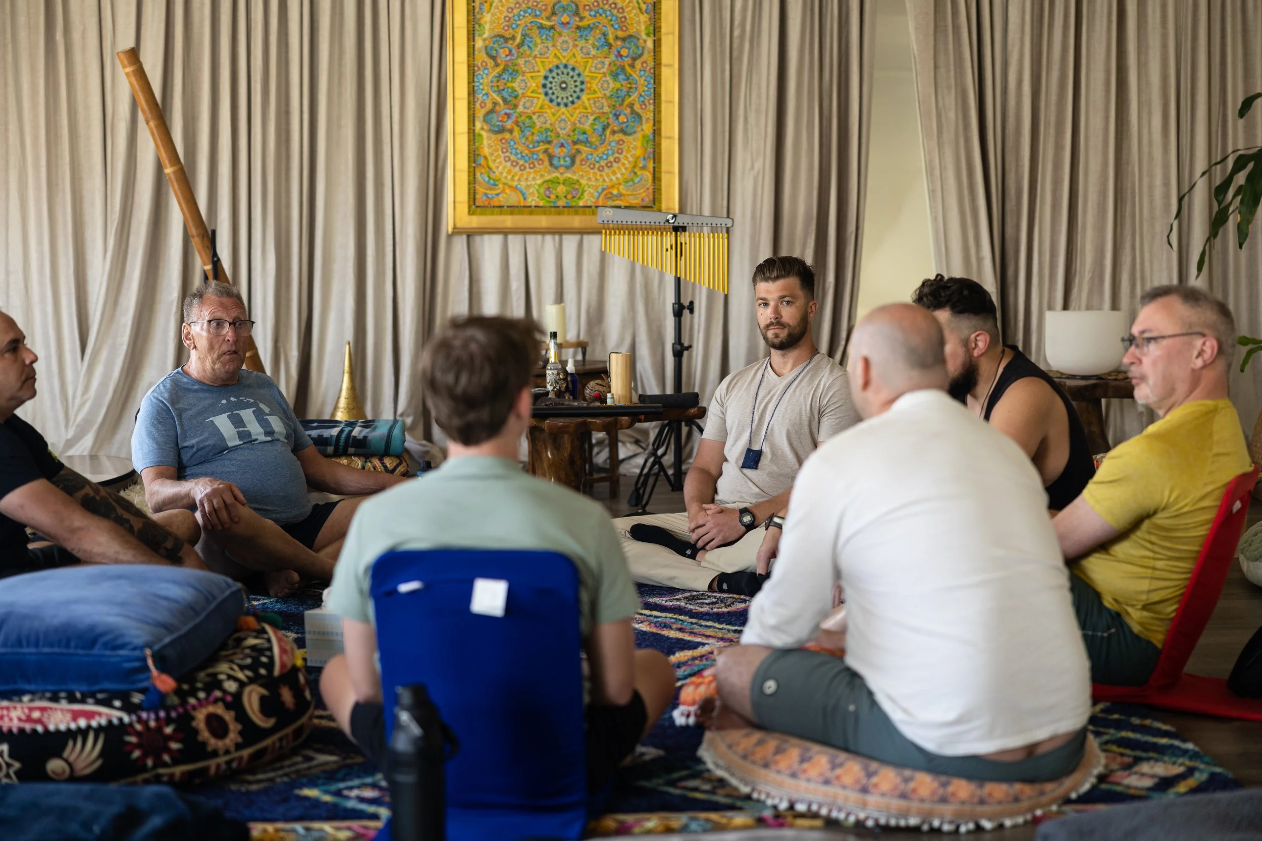 A group of people sitting on the floor in a circle during a discussion or meditation session, with musical instruments and colorful wall art in the background.
