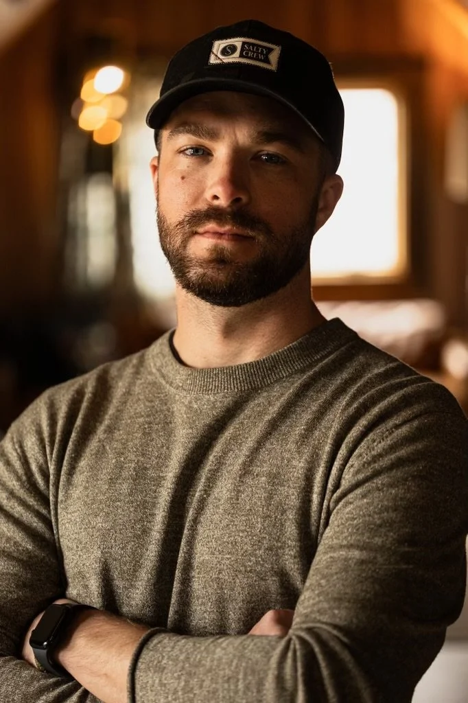 A man with a beard wearing a black cap and a gray sweater, standing indoors with crossed arms.