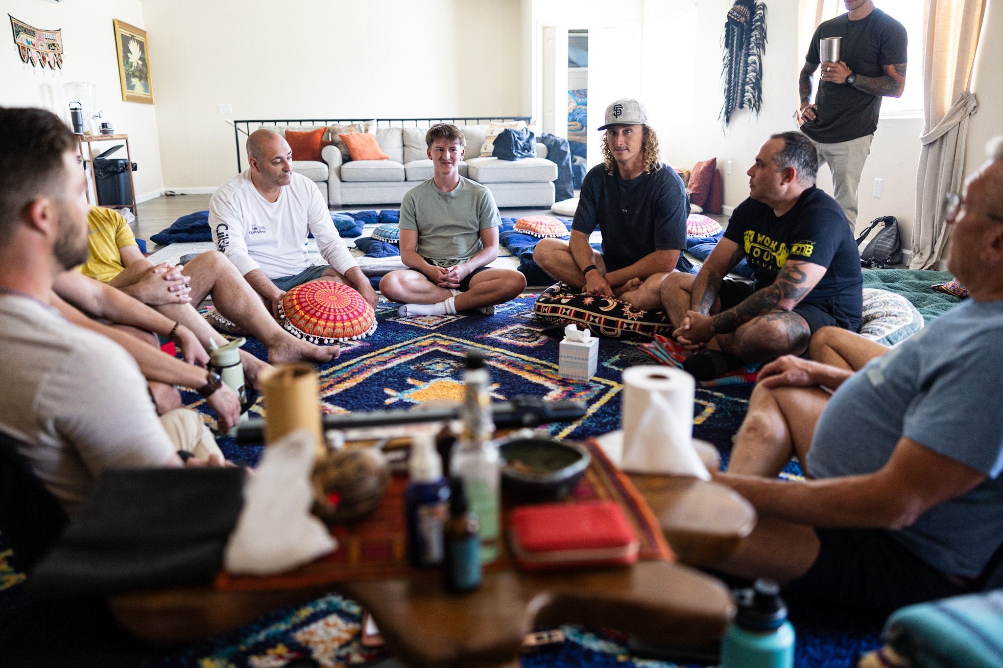 A group of people sitting cross-legged on the floor in a living room, engaged in a discussion or activity. There are cushions and a colorful rug, with various personal bags and items scattered around. A man in a black t-shirt and beige pants is standing to the side, holding a tumbler.