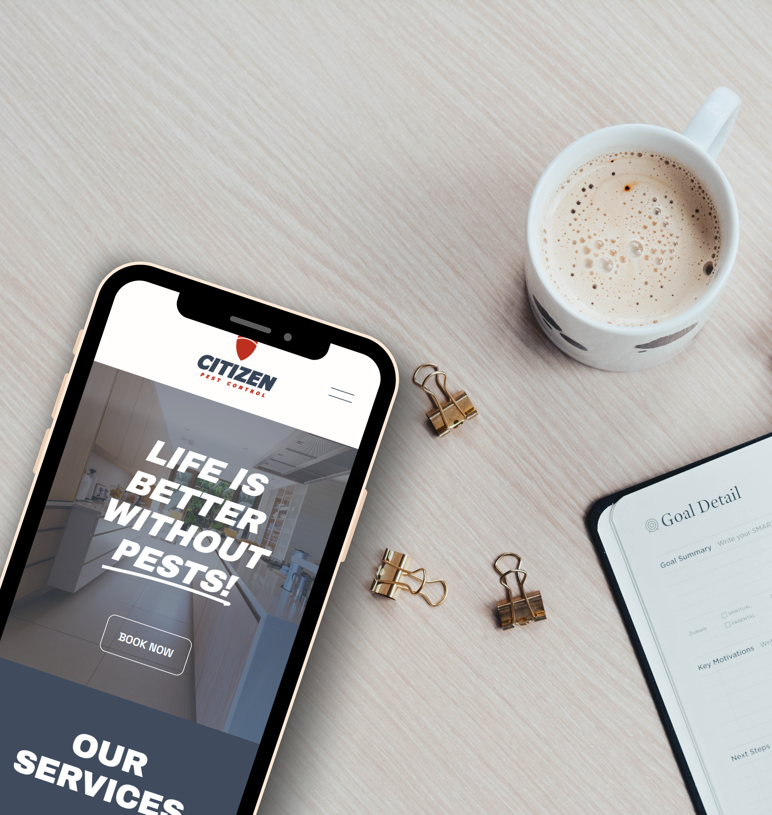 A smartphone displaying a pest control website with the slogan 'Life is better without pests!', a notebook titled 'Goal Detail,' a cup of coffee or tea, and three gold binder clips on a light wooden desk.