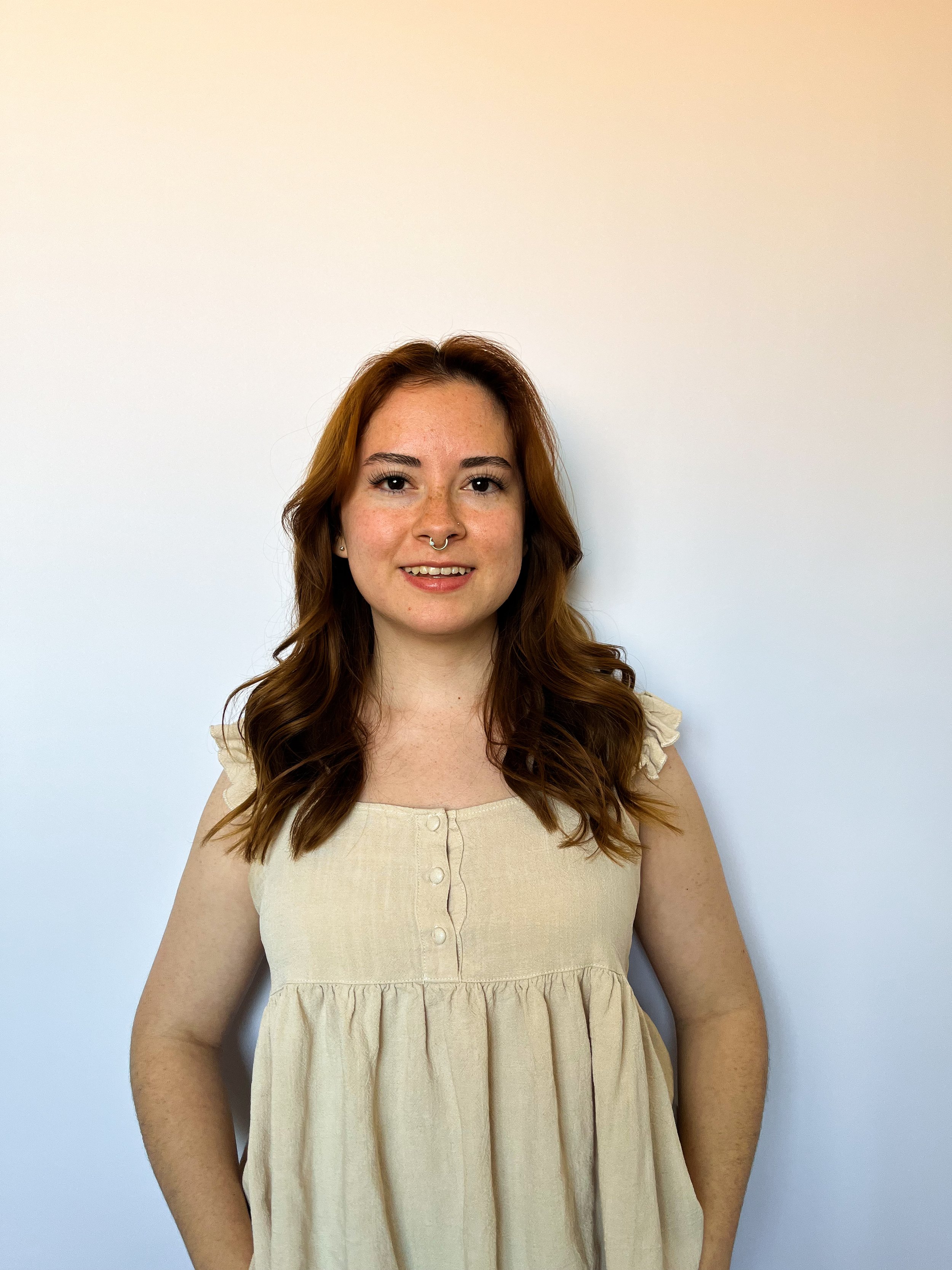 A young woman with red hair, freckles, and a septum piercing is standing against a plain white wall, smiling at the camera.