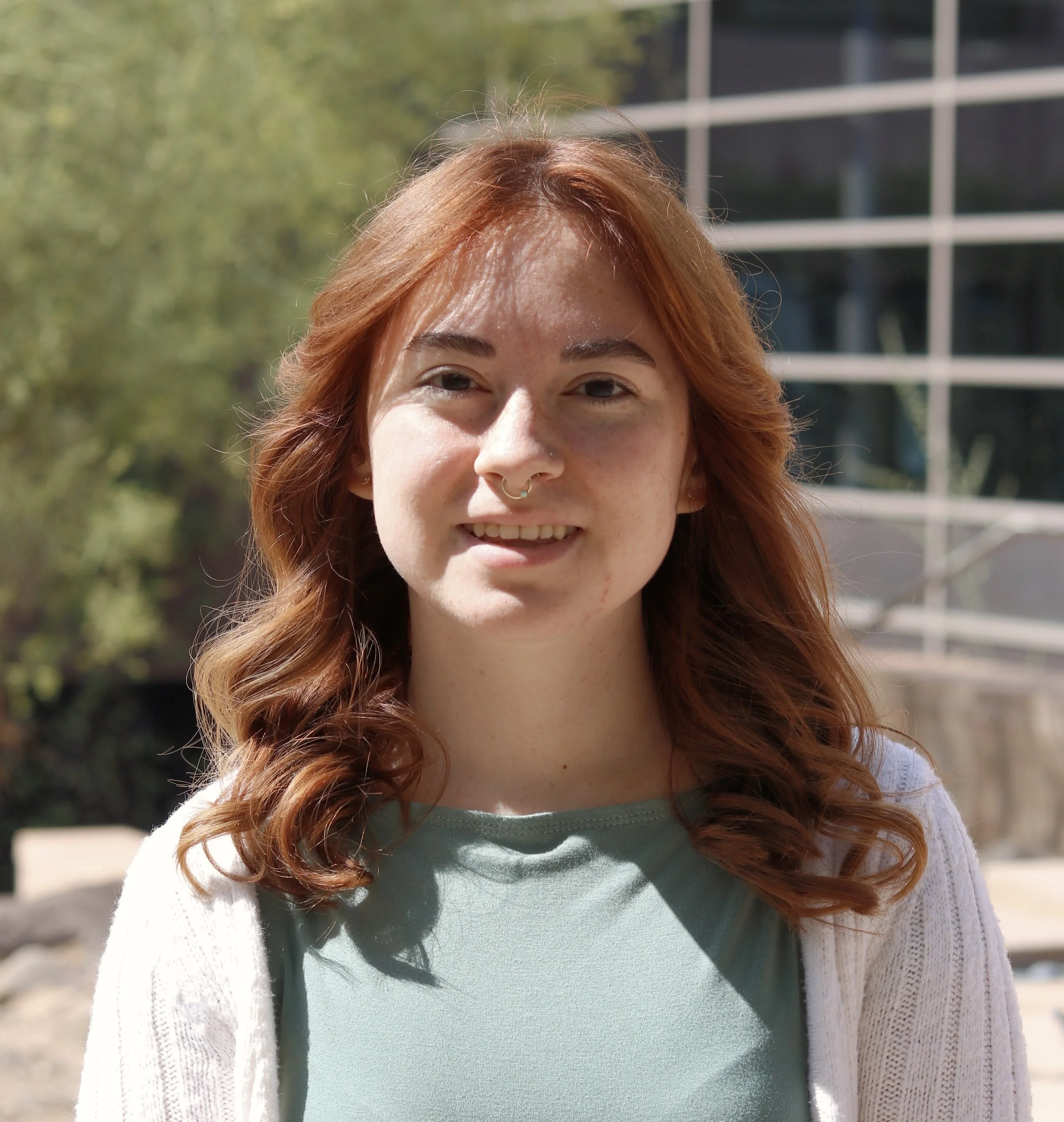 A headshot of the founder of Cornerstone Collective LLC, young woman with wavy red hair and a septum piercing smiling outdoors on a sunny day, with a blurred modern building and trees in the background.