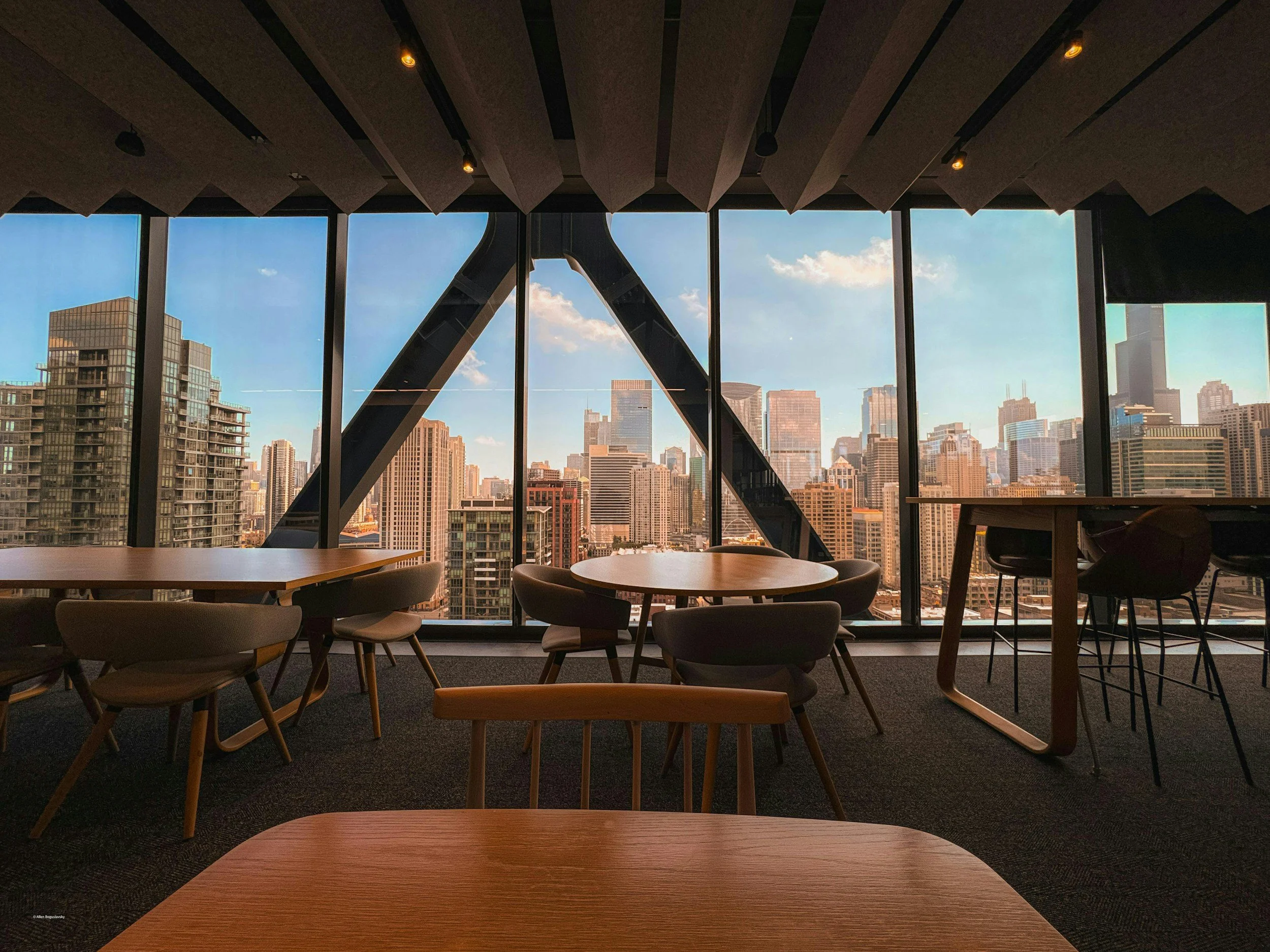 Interior of a modern office with round and rectangular tables, chairs, and large floor-to-ceiling windows showcasing a city skyline with tall buildings and blue sky.