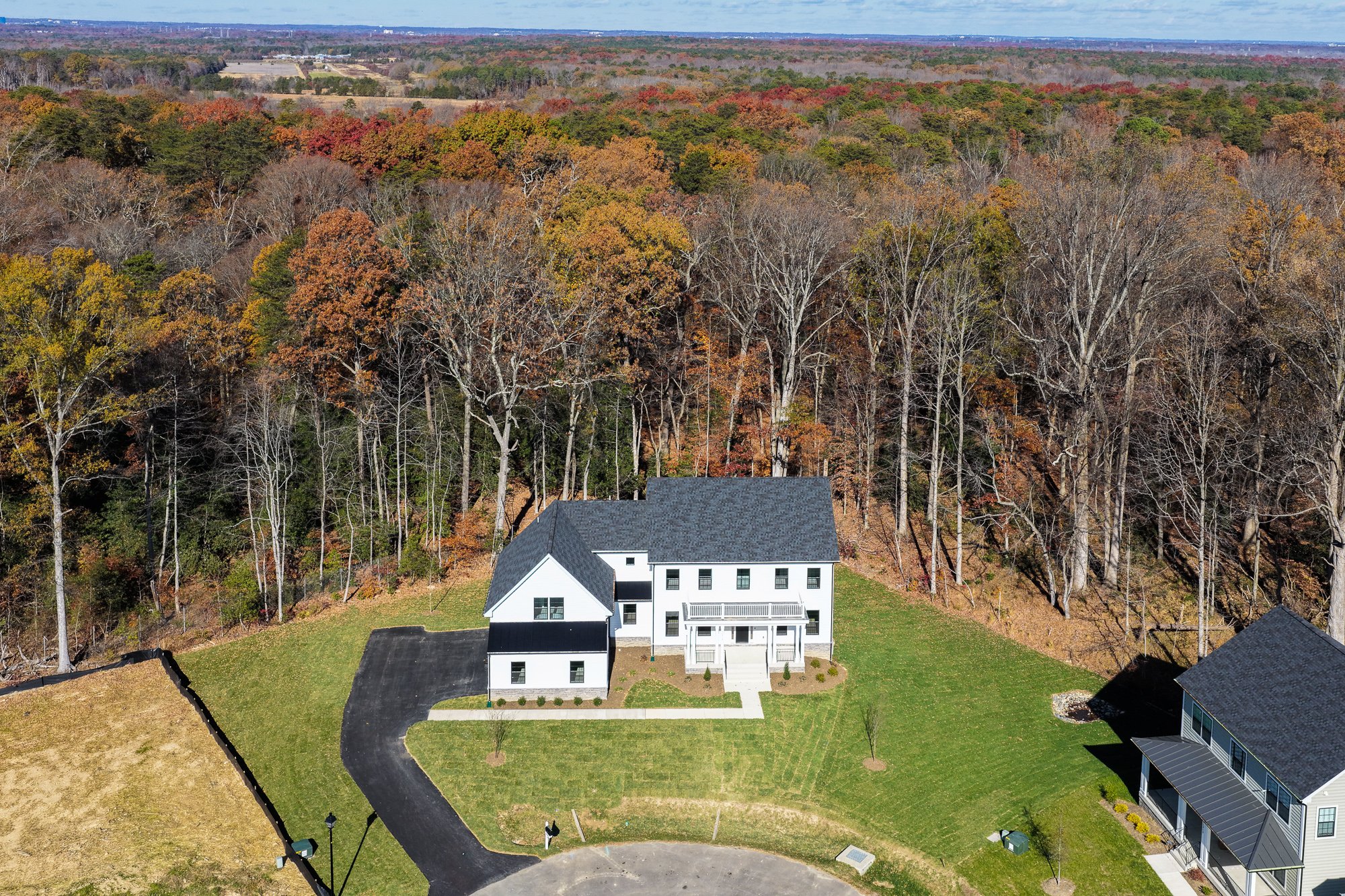 Aerial view of a new white house with black roof and attached garage, surrounded by a well-manicured lawn, with neighboring house to the right, and a dense forest in the background.