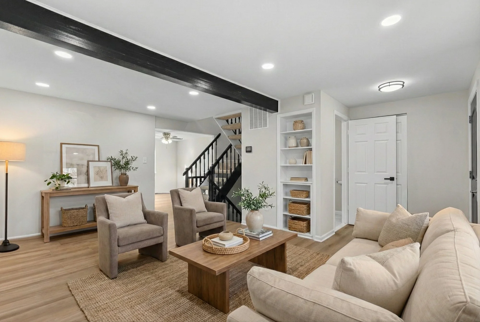 A cozy living room with beige sofas and armchairs, a wooden coffee table, and a white bookshelf with decorative items; wood flooring, a black staircase, and neutral-colored walls.