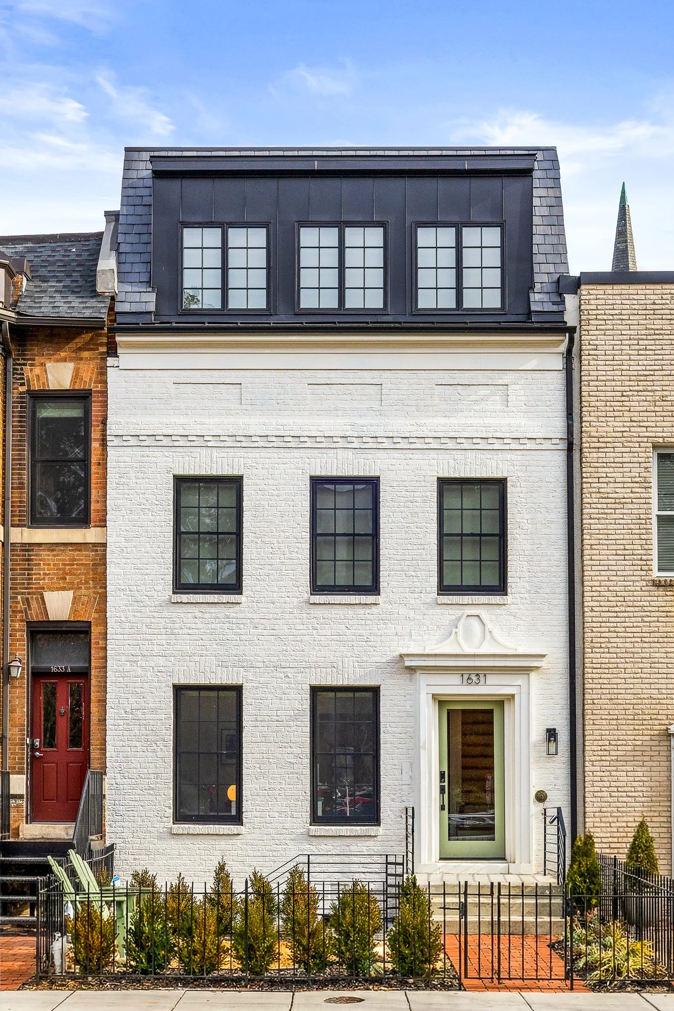 A white brick townhouse with black-framed windows and a light green front door, surrounded by a small garden with shrubs and a black fence.