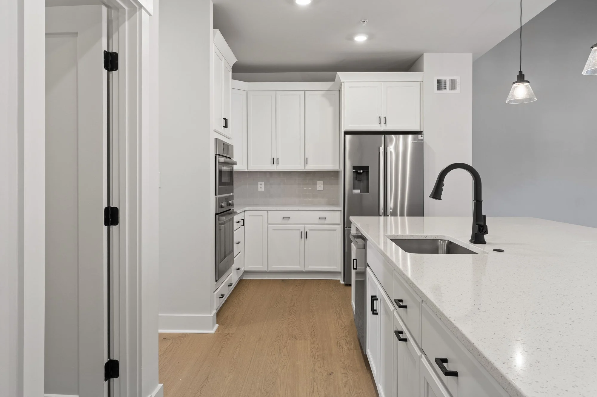 Modern kitchen with white cabinets, stainless steel refrigerator, double oven, black faucet, white countertop, and light wood flooring.