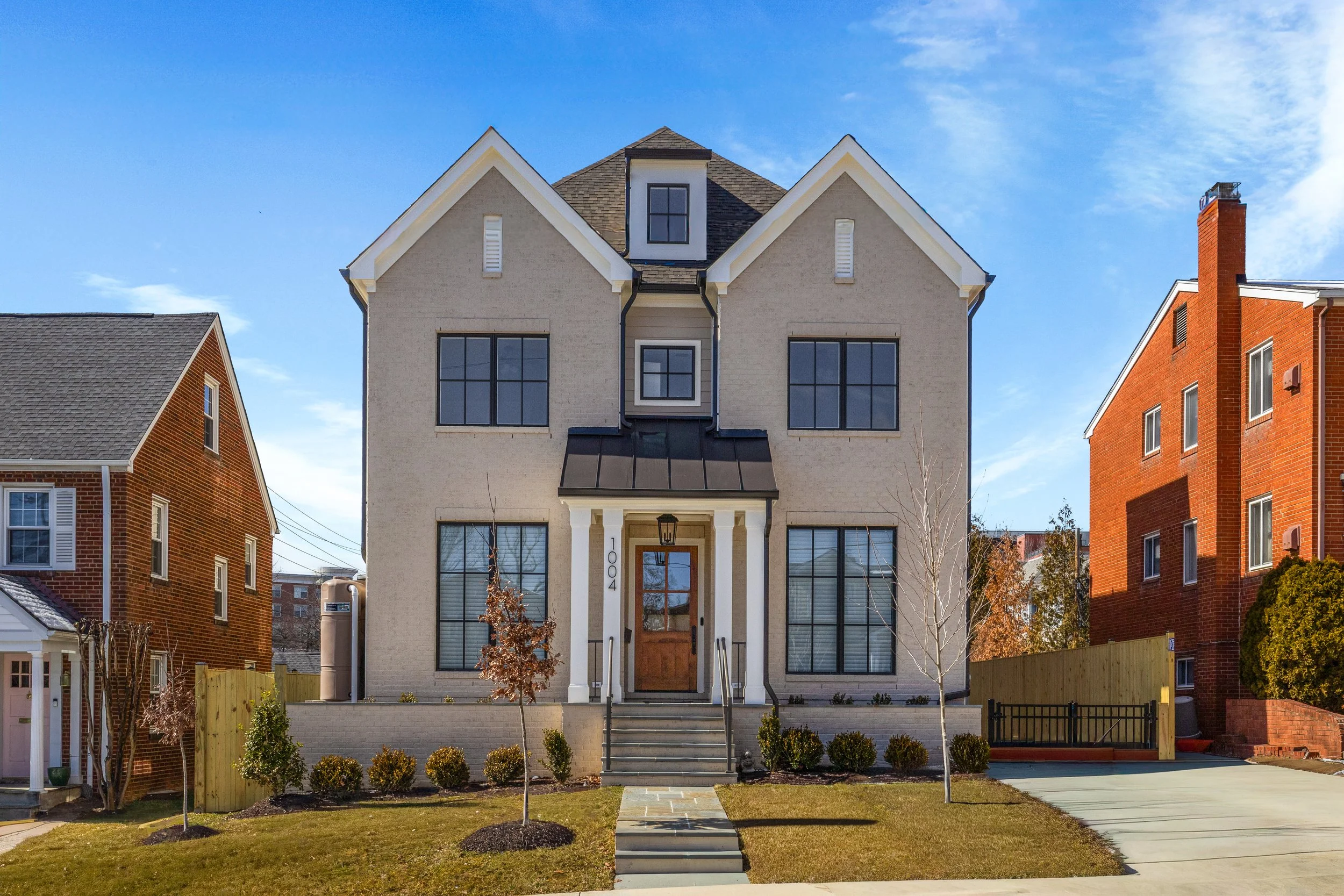 Front view of a modern, light-colored house with a gabled roof, black-framed windows, and a wooden front door, flanked by small trees and bushes, with neighboring brick houses on both sides, under a clear blue sky.