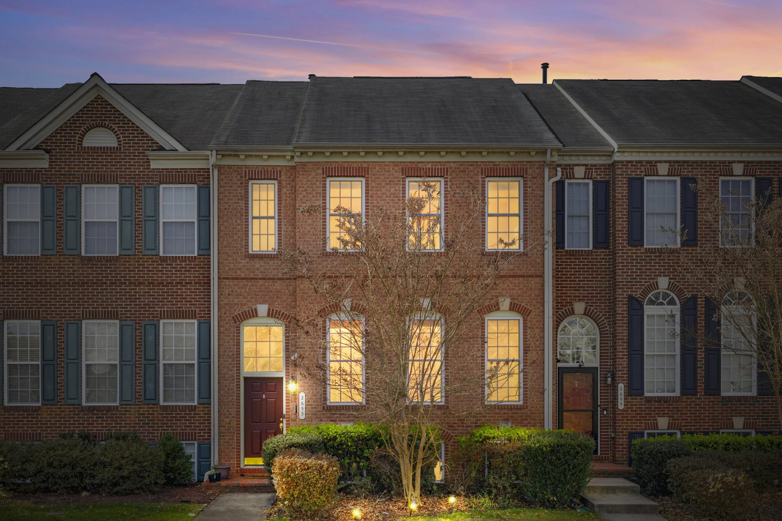 Row of townhouse apartments at sunset with lit windows and a tree in front.