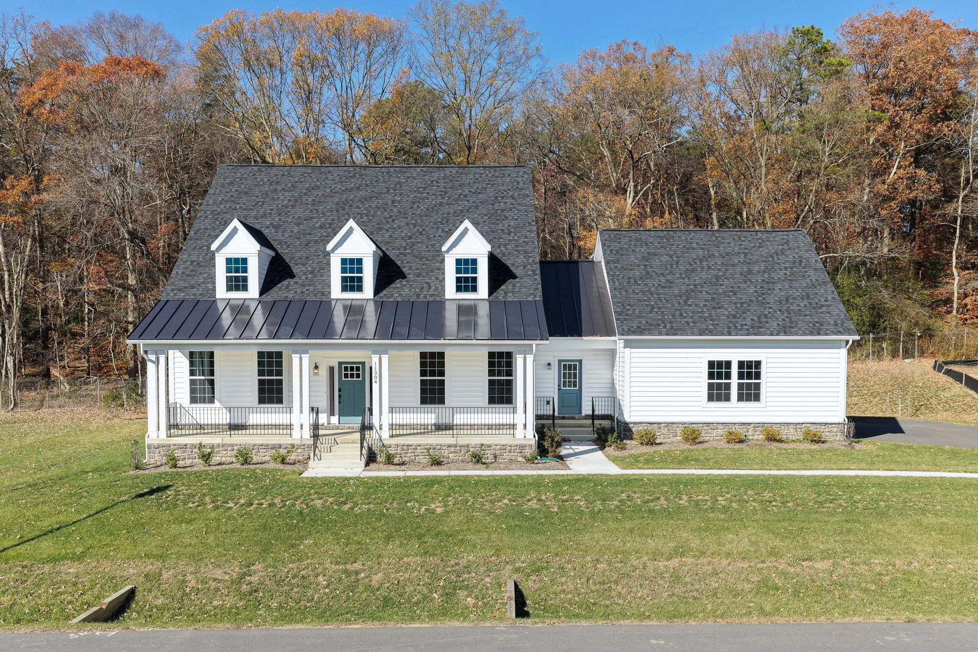 A white house with black roof shingles, three dormer windows, a front porch with black railing, and a small backyard with a grassy area. Trees with fall foliage are in the background.