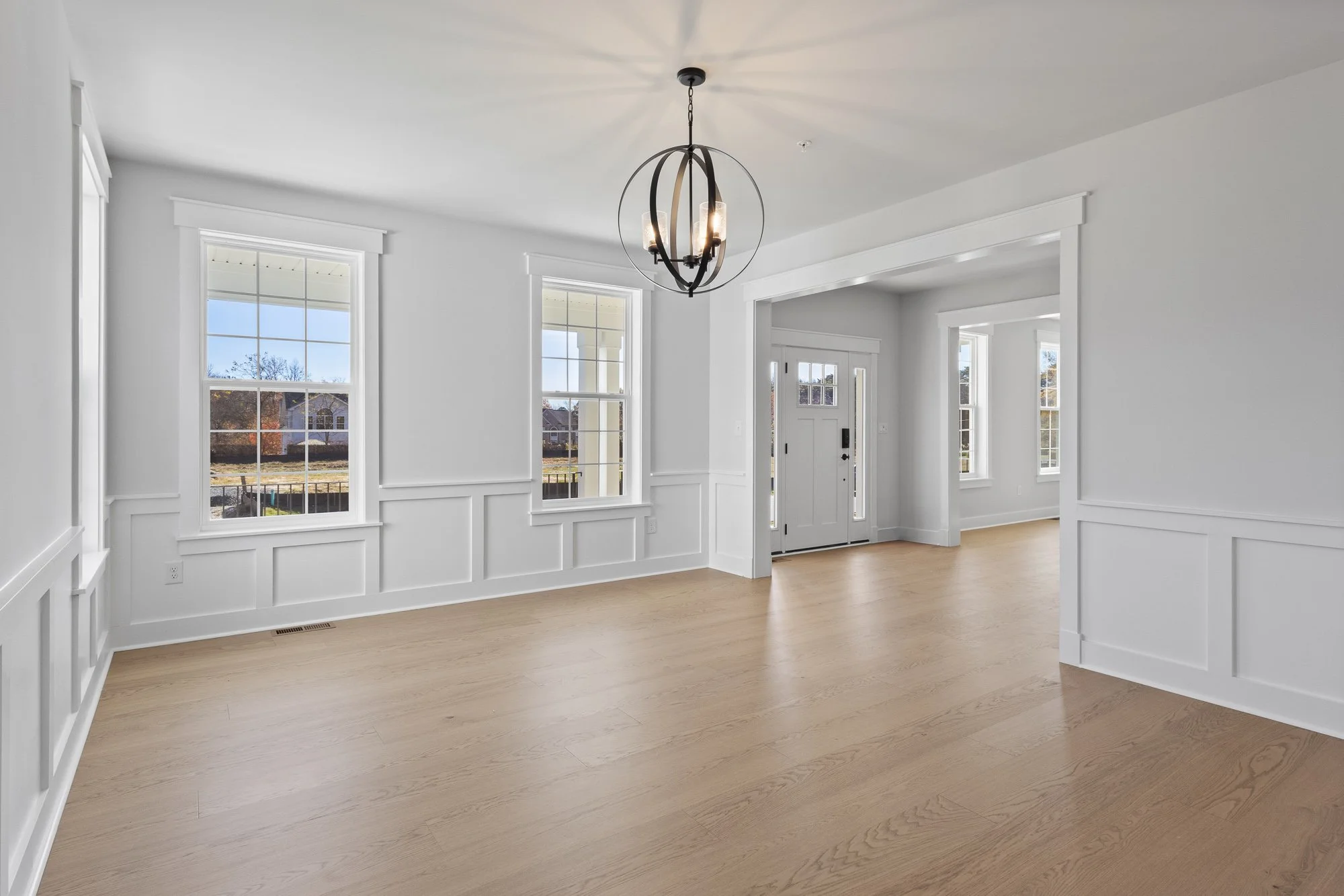 Empty living room with white walls, large windows, light wood flooring, and a modern chandelier hanging from the ceiling.