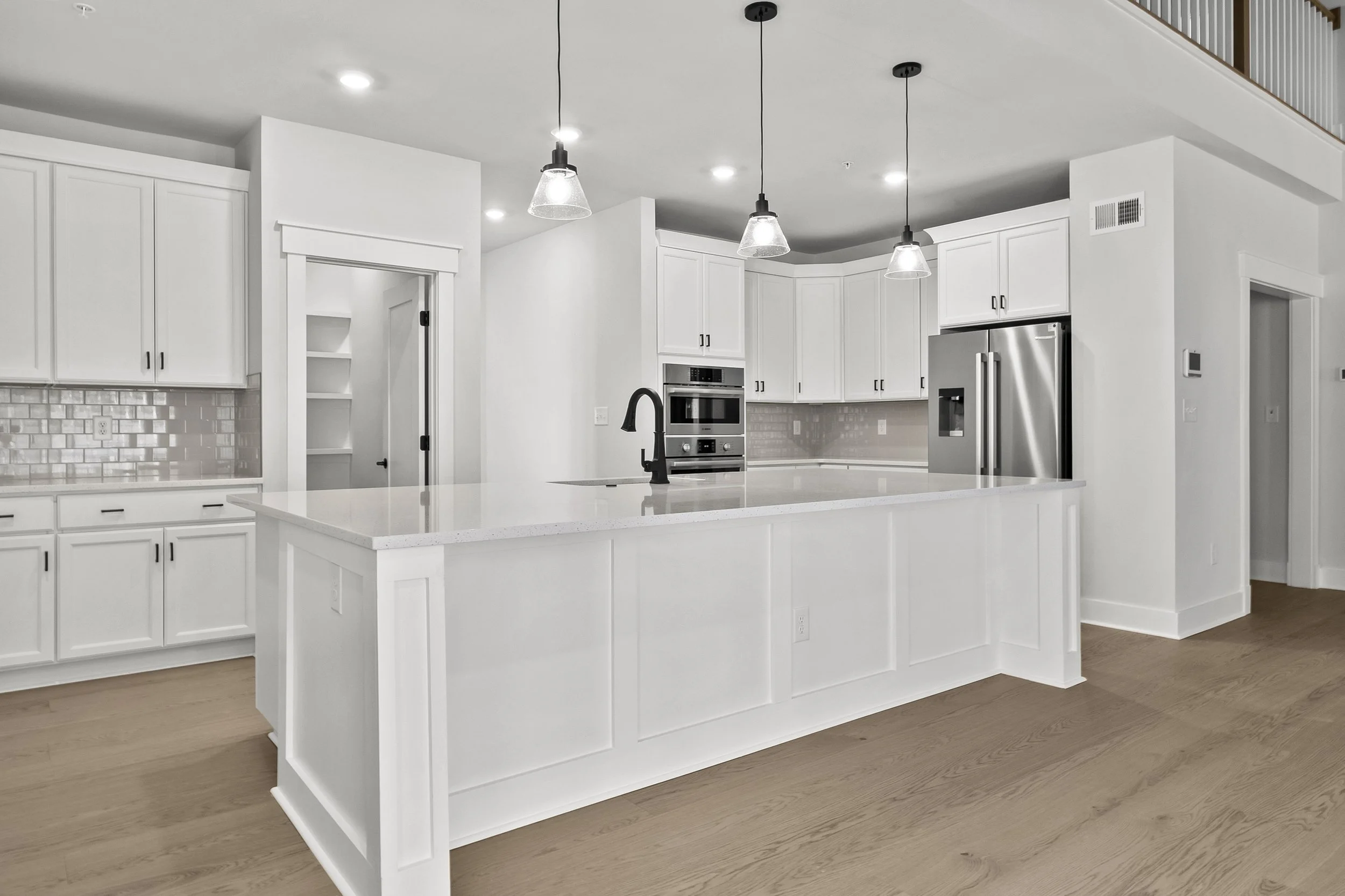 A modern kitchen in Glenn Dale, MD with white cabinets, a large island with a white countertop, black fixtures, pendant lights, stainless steel refrigerator, oven, and microwave, and light wood flooring.