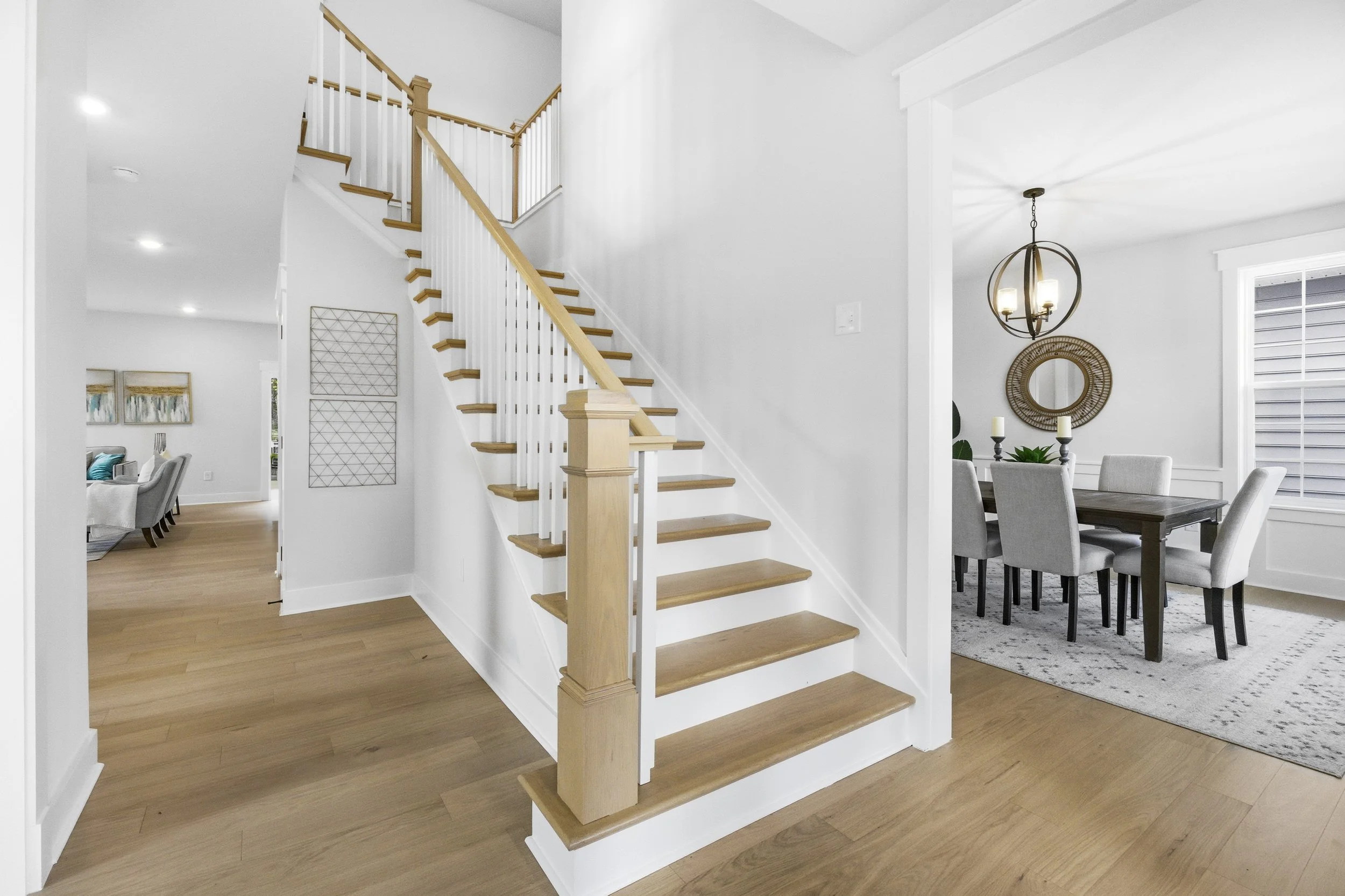 Interior view of a modern home with wooden stairs, white walls, and a dining area with a dark table and light chairs.