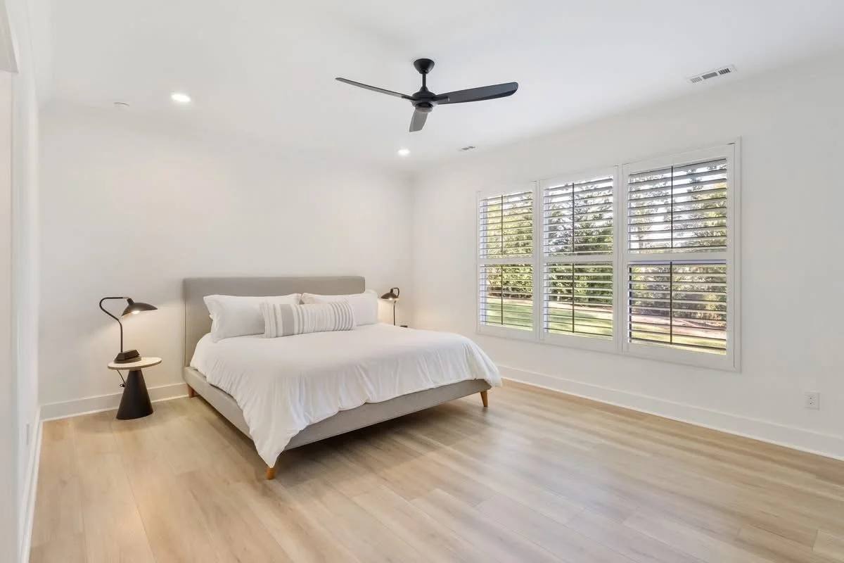 Empty bedroom with white walls, light wood flooring, large windows with white shutters, a ceiling fan, and a bed with white bedding and minimalist black bedside lamps.