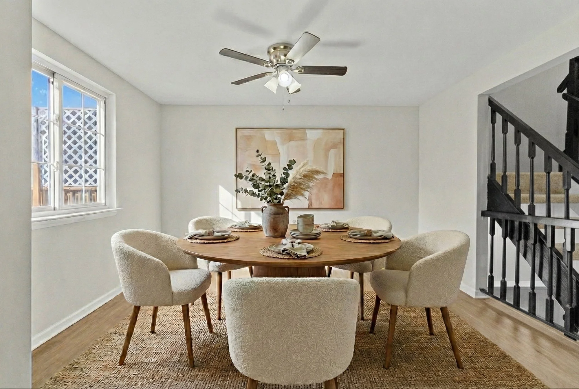A dining room with a round wooden table set with plates, bowls, napkins, and utensils, surrounded by six upholstered chairs, a centerpiece with greenery and pampas grass, a window with lattice outside, a painting on the wall, a ceiling fan, and a staircase with black railing.