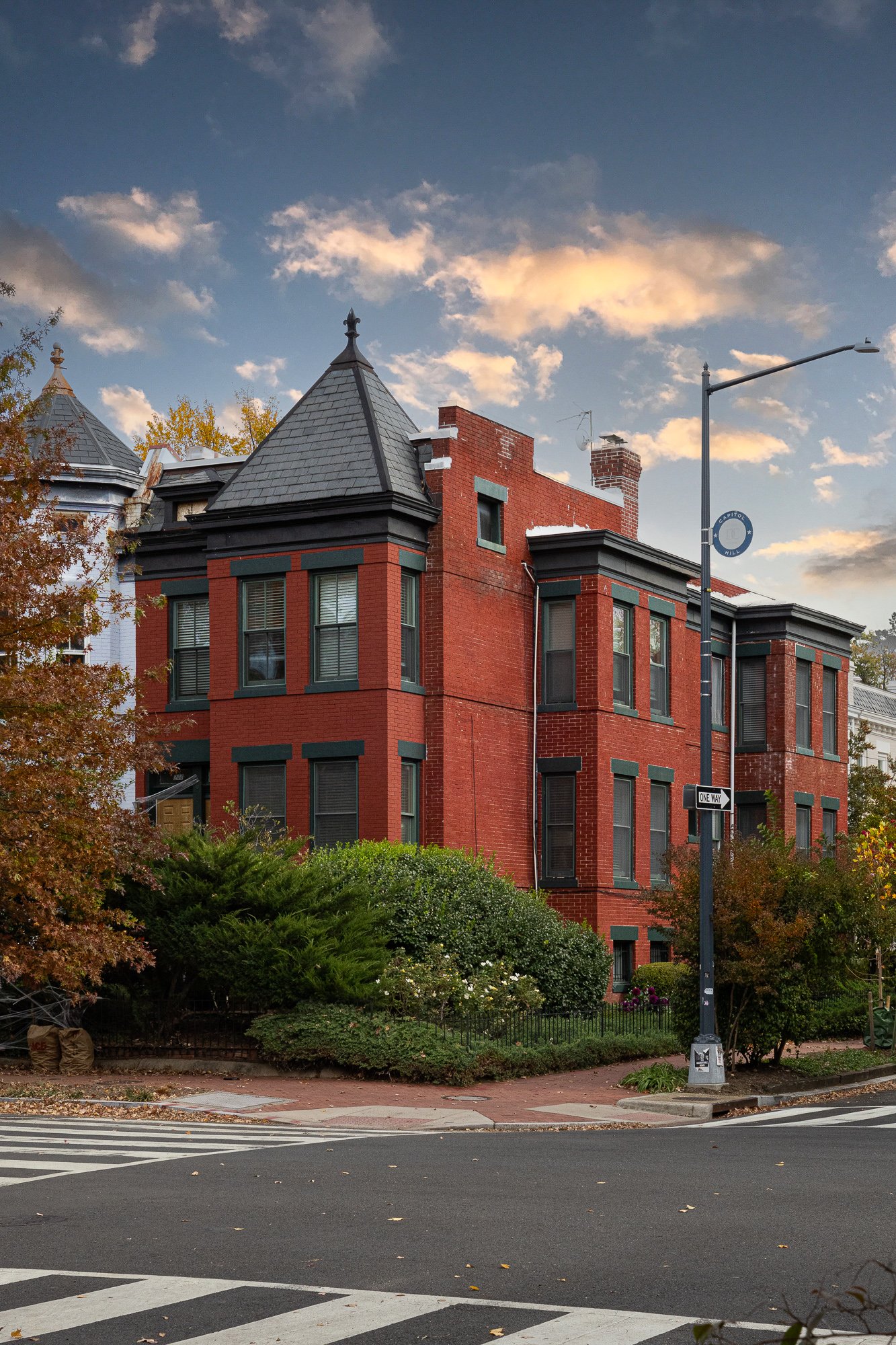 A red brick Victorian-style house with dark trim, turrets, and bay windows on a city street with crosswalk and street signs, trees with autumn leaves, and a partly cloudy sky.