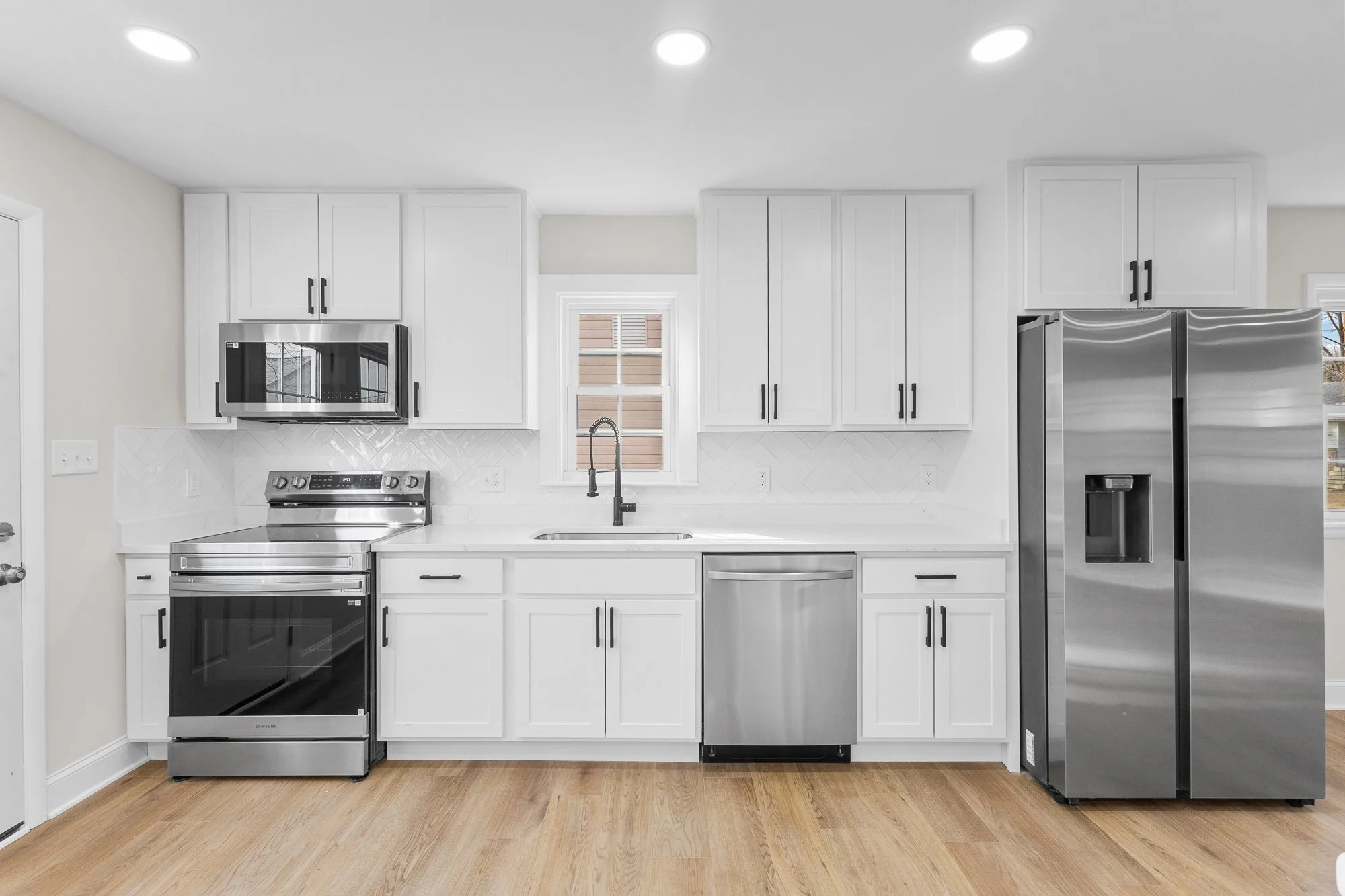 Modern kitchen with white cabinets, stainless steel appliances including a fridge, oven, microwave, and dishwasher, farmhouse sink, wooden flooring, and window above the sink.