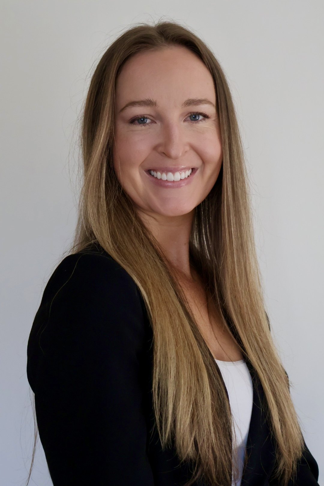 A woman with long light brown hair, wearing a black blazer and white top, smiling against a plain light-colored background.