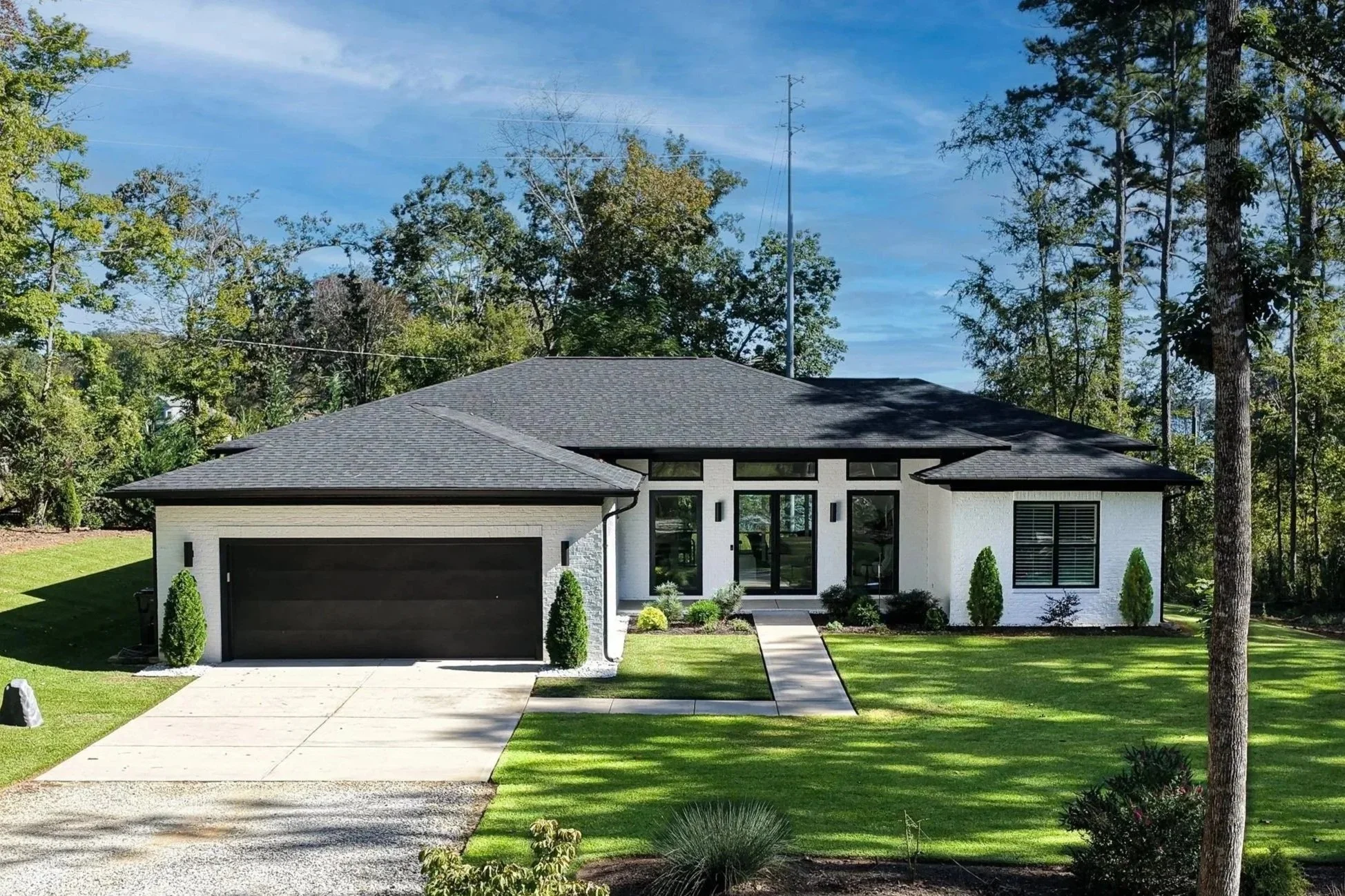 Modern single-story house with white exterior walls, black garage door, and black window frames, surrounded by green lawn and trees under a blue sky.