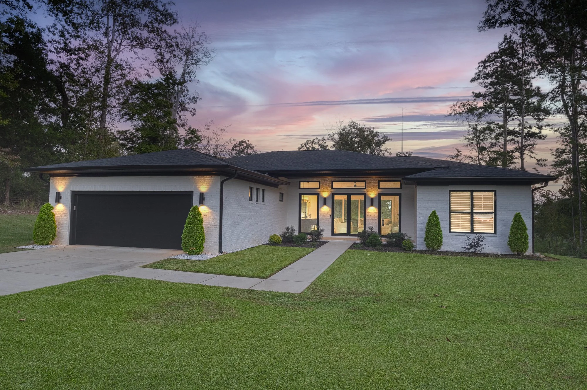 Modern single-story house with white brick exterior, black window frames, and a black garage door. The house is illuminated by exterior lights, with a neatly maintained lawn and small bushes along the front walkway. The sky in the background shows a colorful sunset with pink and purple hues.