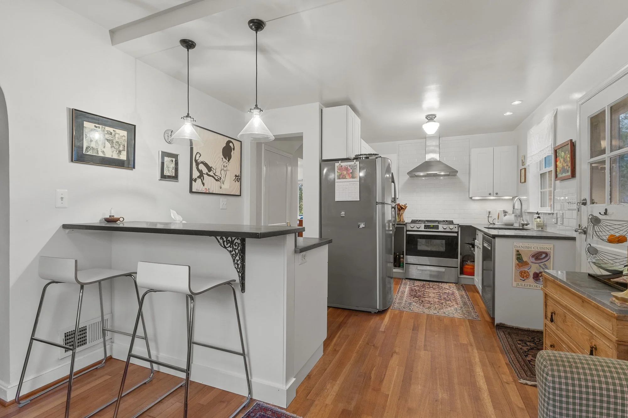 A kitchen with white cabinets, a stainless steel refrigerator, a stove, and a range hood. There are two pendant lights hanging above a breakfast bar with two white chairs. The kitchen floor is wooden, and there are framed pictures on the walls and a window with blinds.