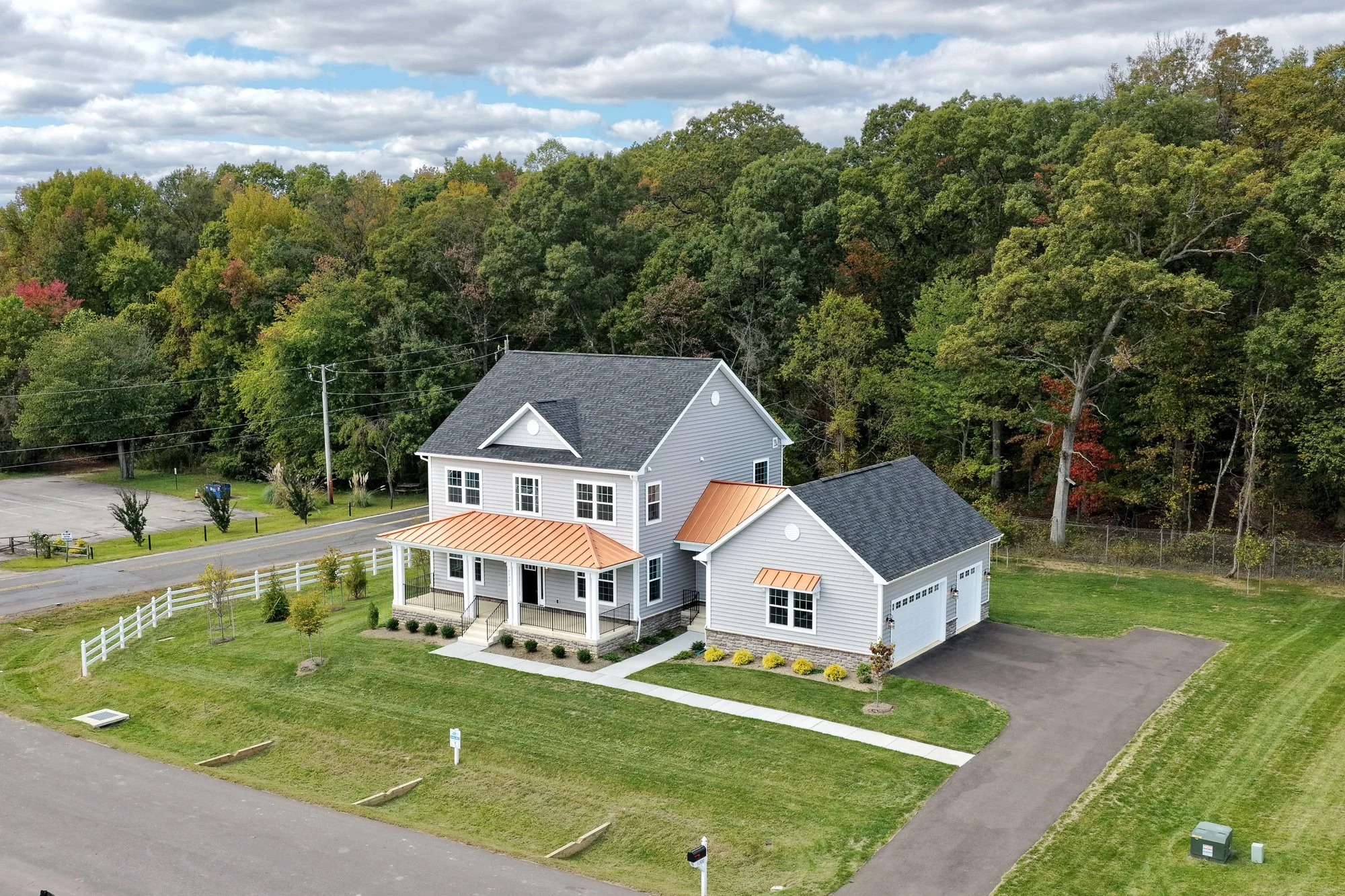 A modern two-story house with gray siding, black roof, and copper accents on the porch and side awnings, surrounded by a well-maintained lawn, small trees, and a white picket fence, with a driveway leading to a garage, neighboring wooded area, and a cloudy sky in the background.
