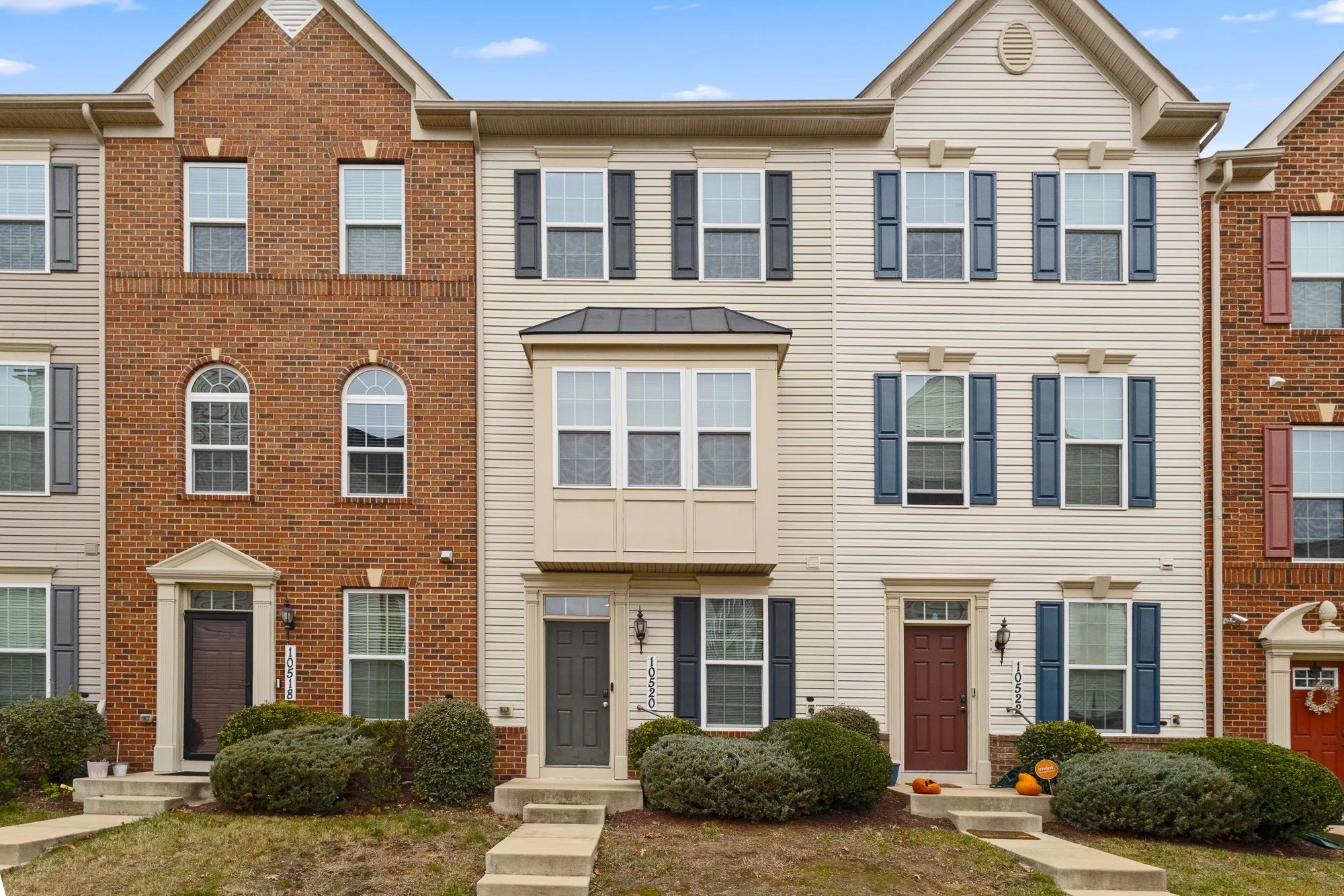 Front view of a multi-story townhouse with a beige vinyl siding facade, black shutters, and a gray front door, flanked by bushes and small steps leading to the entrance.