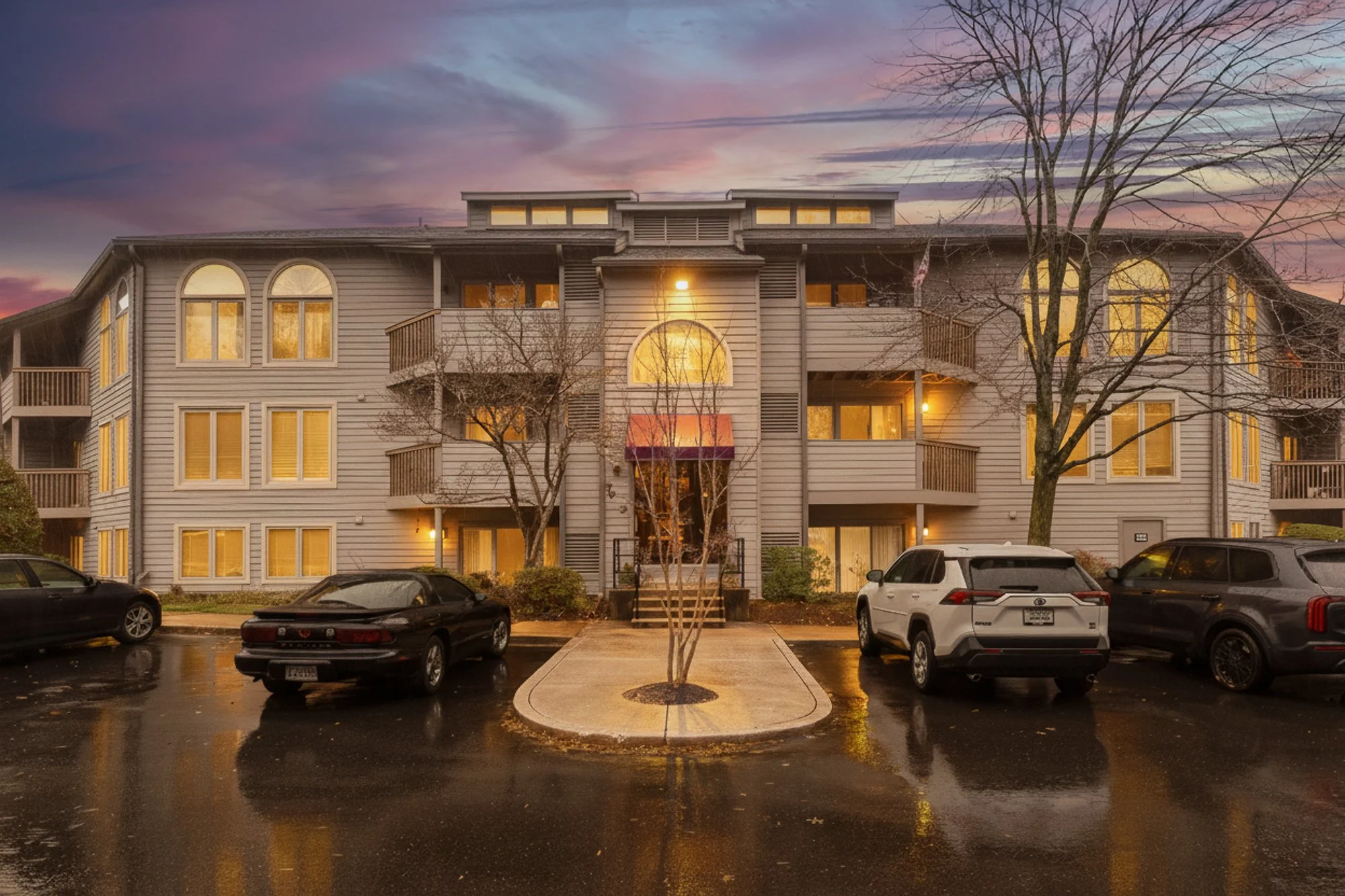 A three-story apartment building with lit windows at sunset, wet parking lot with four parked cars, leafless trees, and a cloudy sky with pink and purple hues.