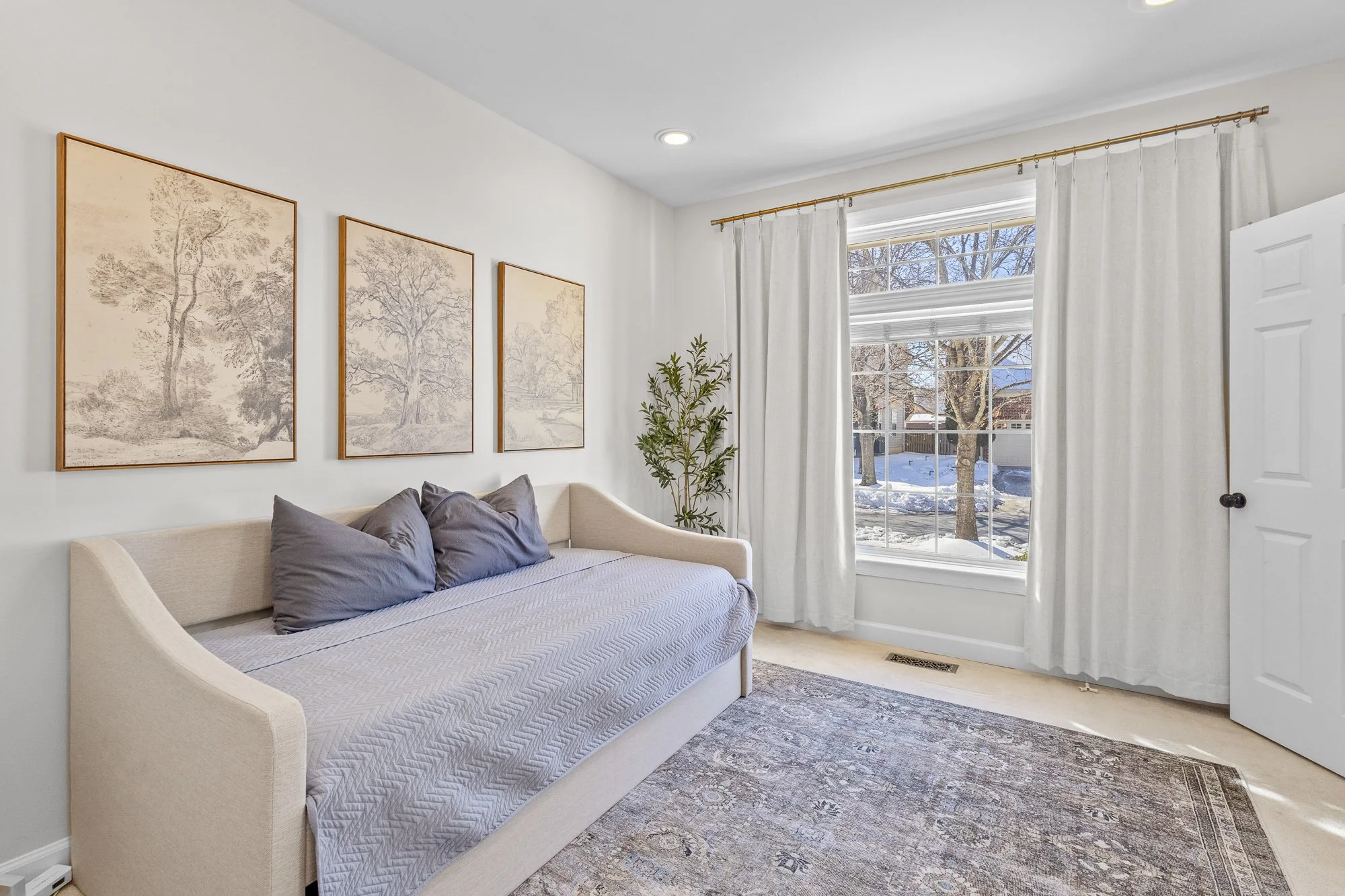 A cozy bedroom with a large window showing a snowy outdoor scene, white curtains, a beige sofa with purple pillows, three framed sketches of trees on the wall, a tall plant near the window, and a patterned area rug on the floor.