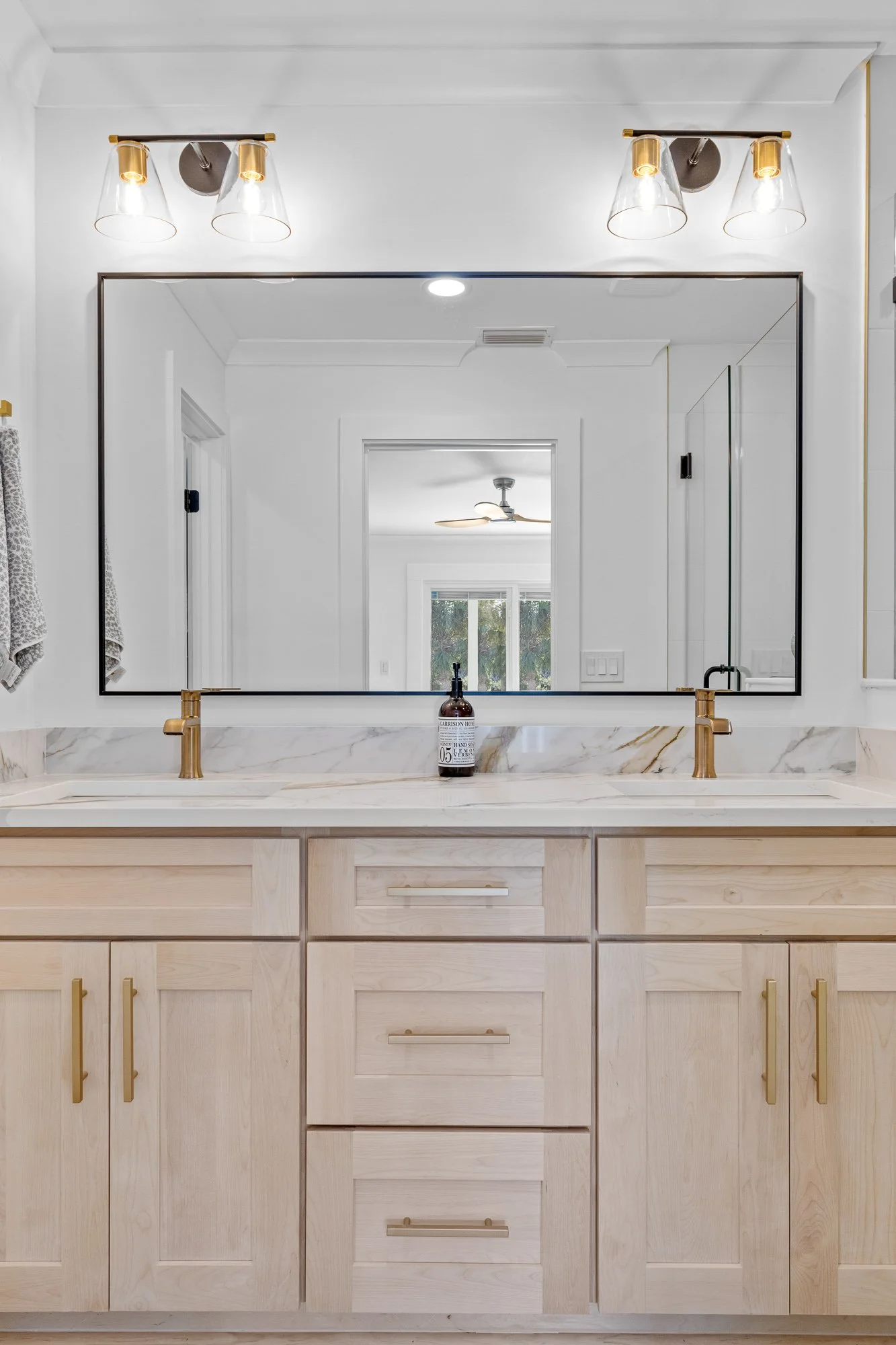 A modern bathroom with a large mirror above a double sink vanity with light wood cabinets. There are two brass faucets and a soap dispenser on the countertop. Light fixtures with glass shades are mounted above the mirror. The reflection shows a ceiling fan and a window in an adjacent room.