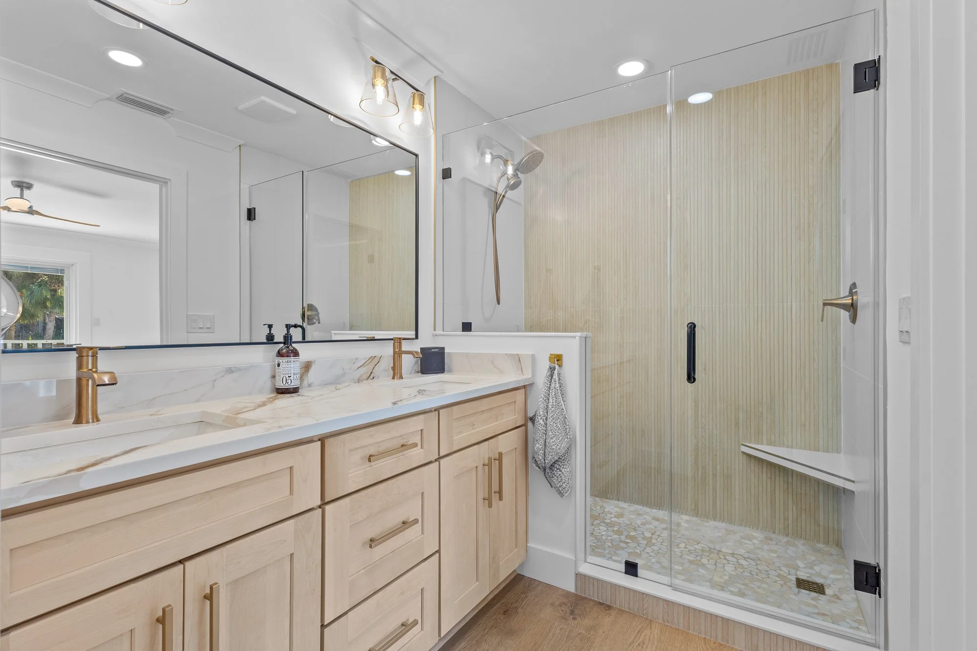 Modern bathroom with wooden vanity, marble countertop, gold fixtures, large mirror, glass-enclosed shower with beige tiled walls, pebble floor, and built-in bench.