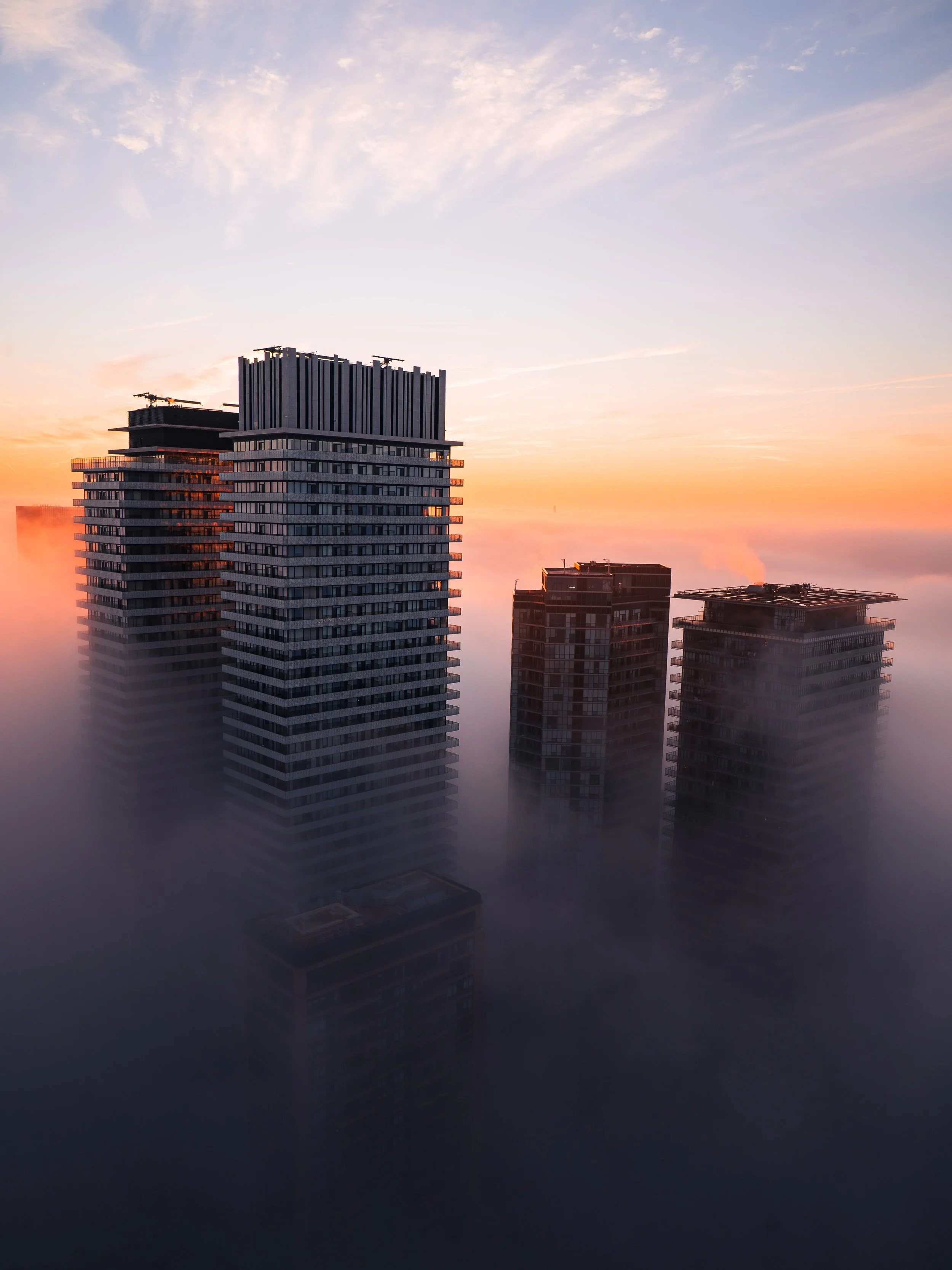 High-rise buildings in a city shrouded in fog with a sunset sky in the background.
