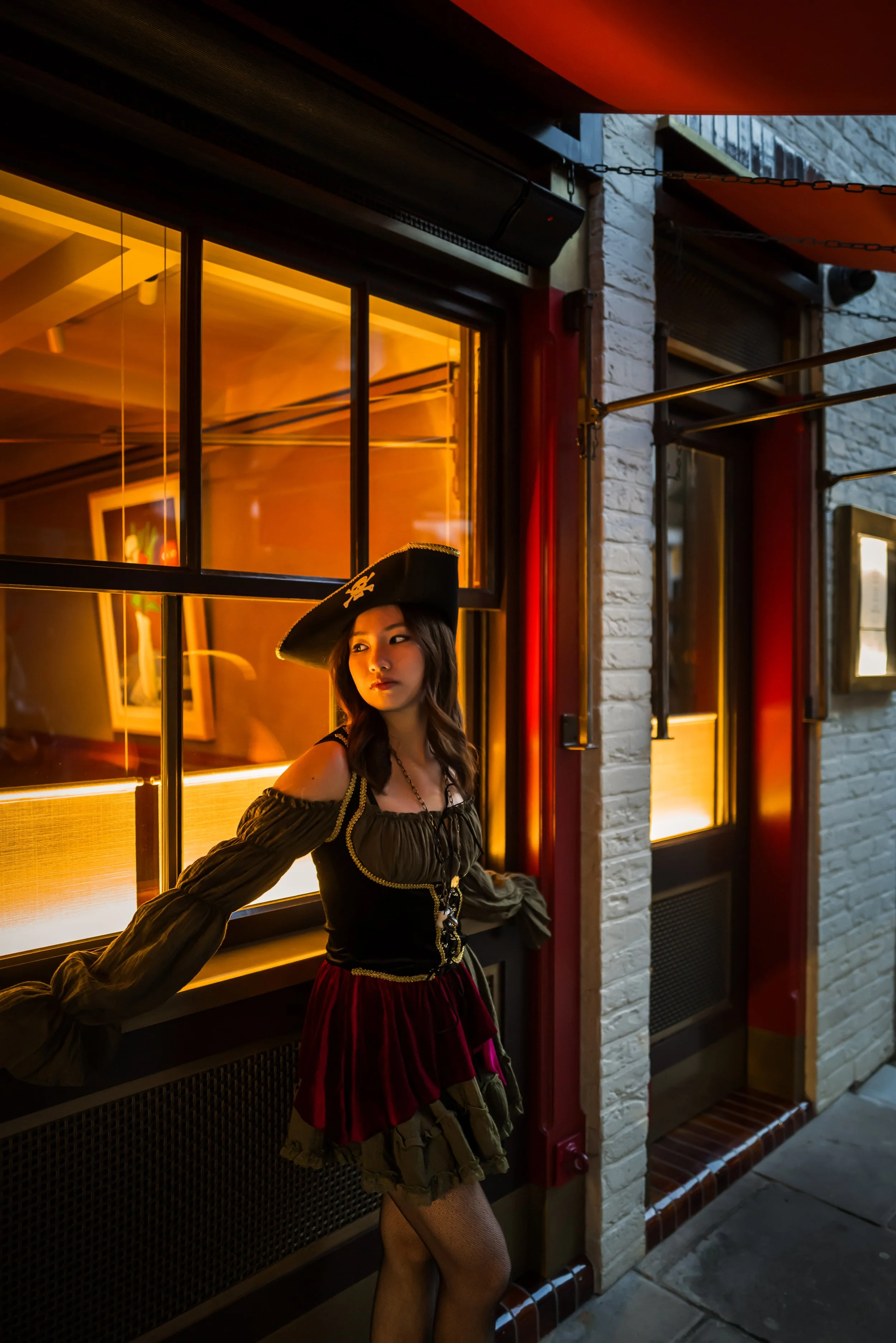 A young woman dressed in a pirate costume with a large hat, black top, red skirt, and fishnet stockings, standing outside a building with brick walls and illuminated windows at night.