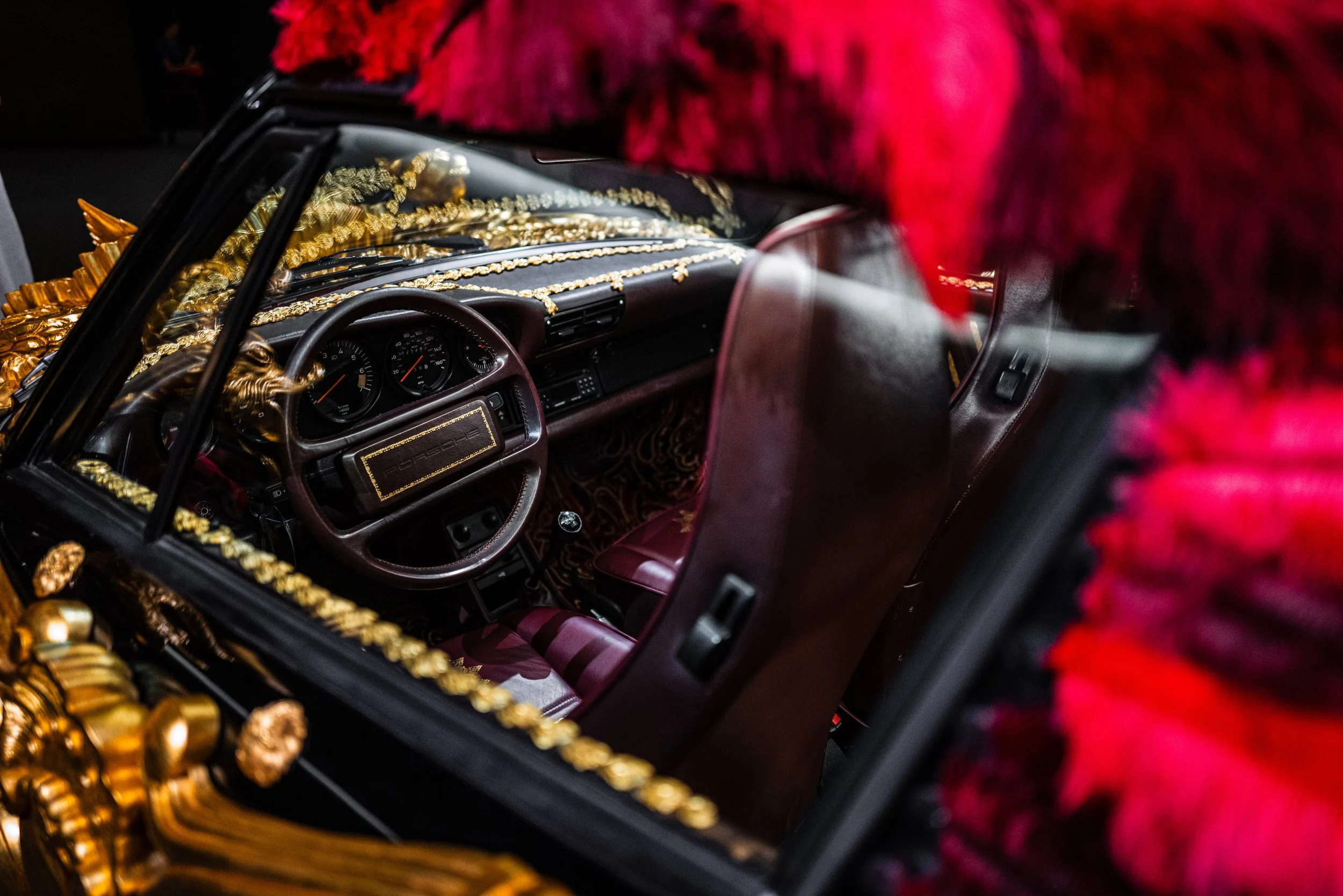 The interior of a luxury car decorated with gold accents, red leather seats, and ornate embellishments, viewed through the window with a partially obscured red and pink fur garment in the foreground.