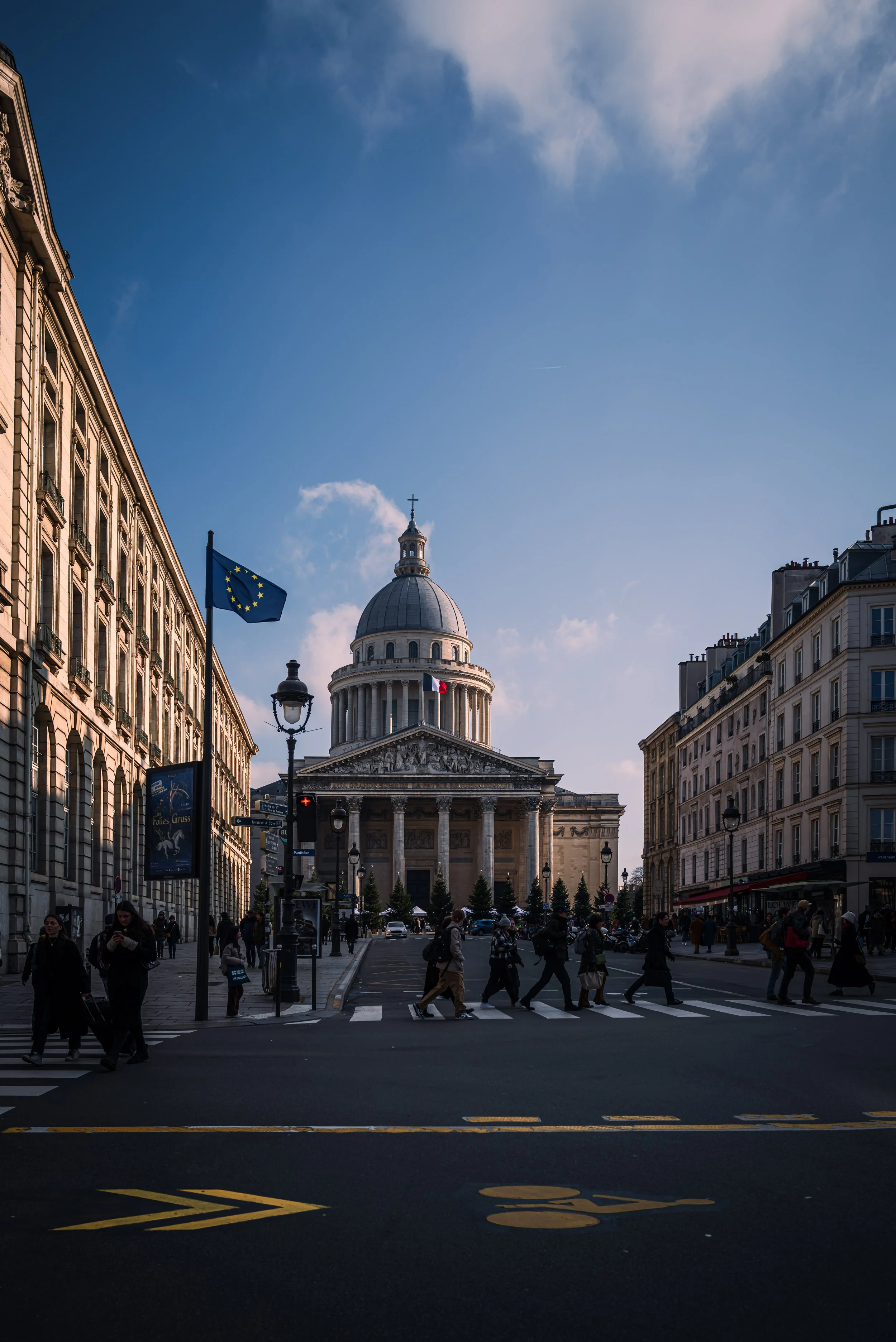 People crossing the street in front of the Panthéon, a historic building in Paris, France, with flags of the European Union and France flying nearby under a blue sky.