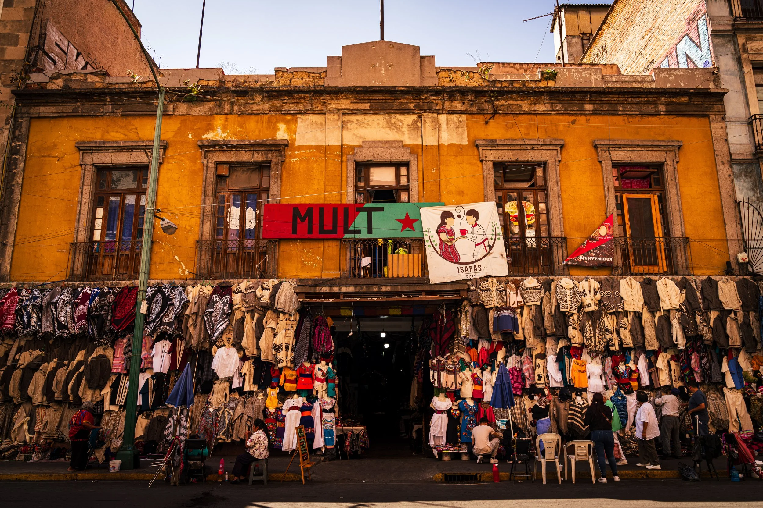 Street scene in Mexico with outdoor clothing and textile stalls in front of a yellow building. The building has two stories with wooden framed windows, and a sign that reads 'MULTI' with a star, and an illustration of two women eating. People are wal