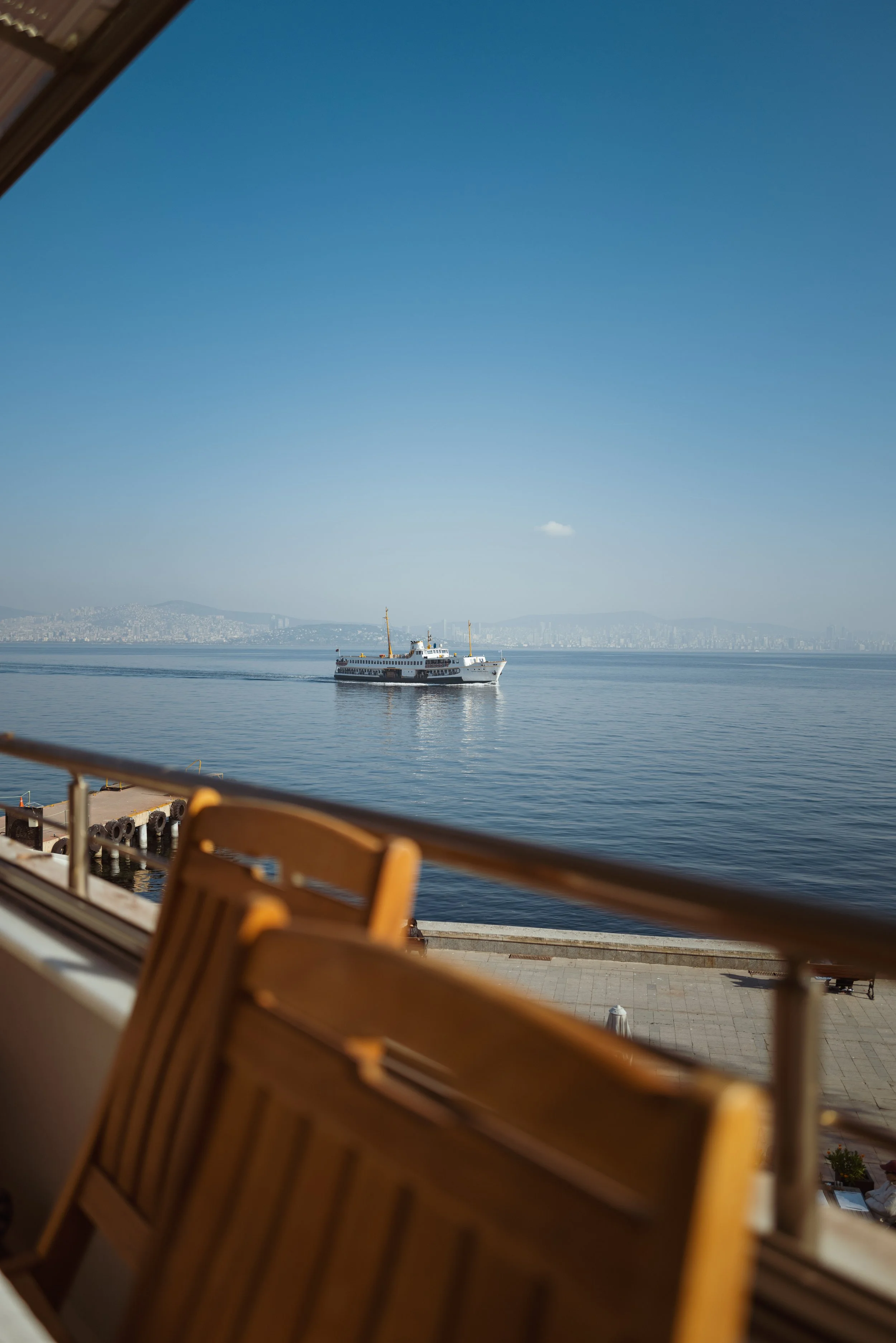 View of a ferry boat sailing on calm water seen from a balcony with wooden chairs, with a city skyline and hills in the background under a clear blue sky.